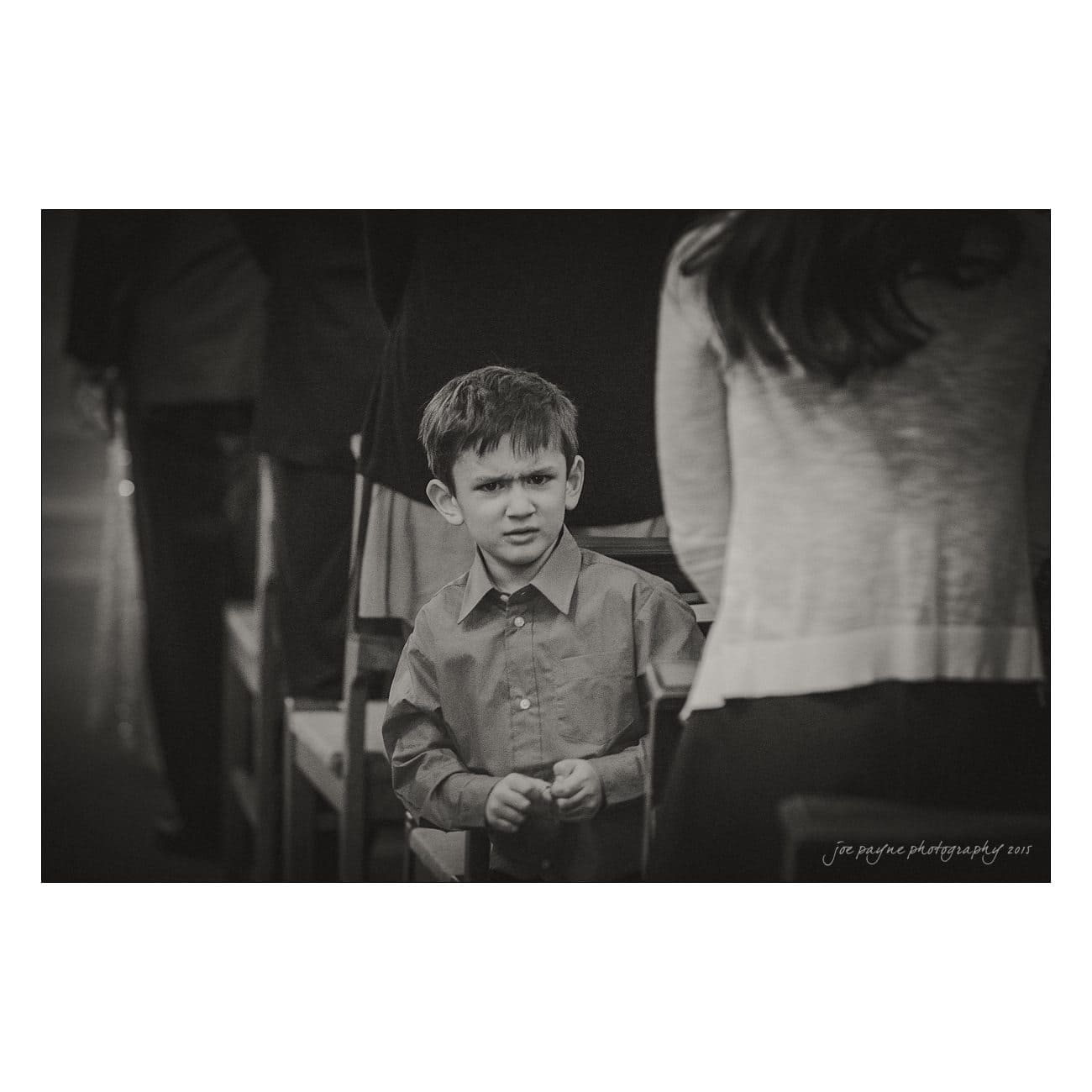 Black and white photo of a young boy standing indoors, looking slightly off to the side with a curious expression. He is surrounded by adults who are partially visible, dressed in formal attire. The photo is signed joe payne photography 2021.
