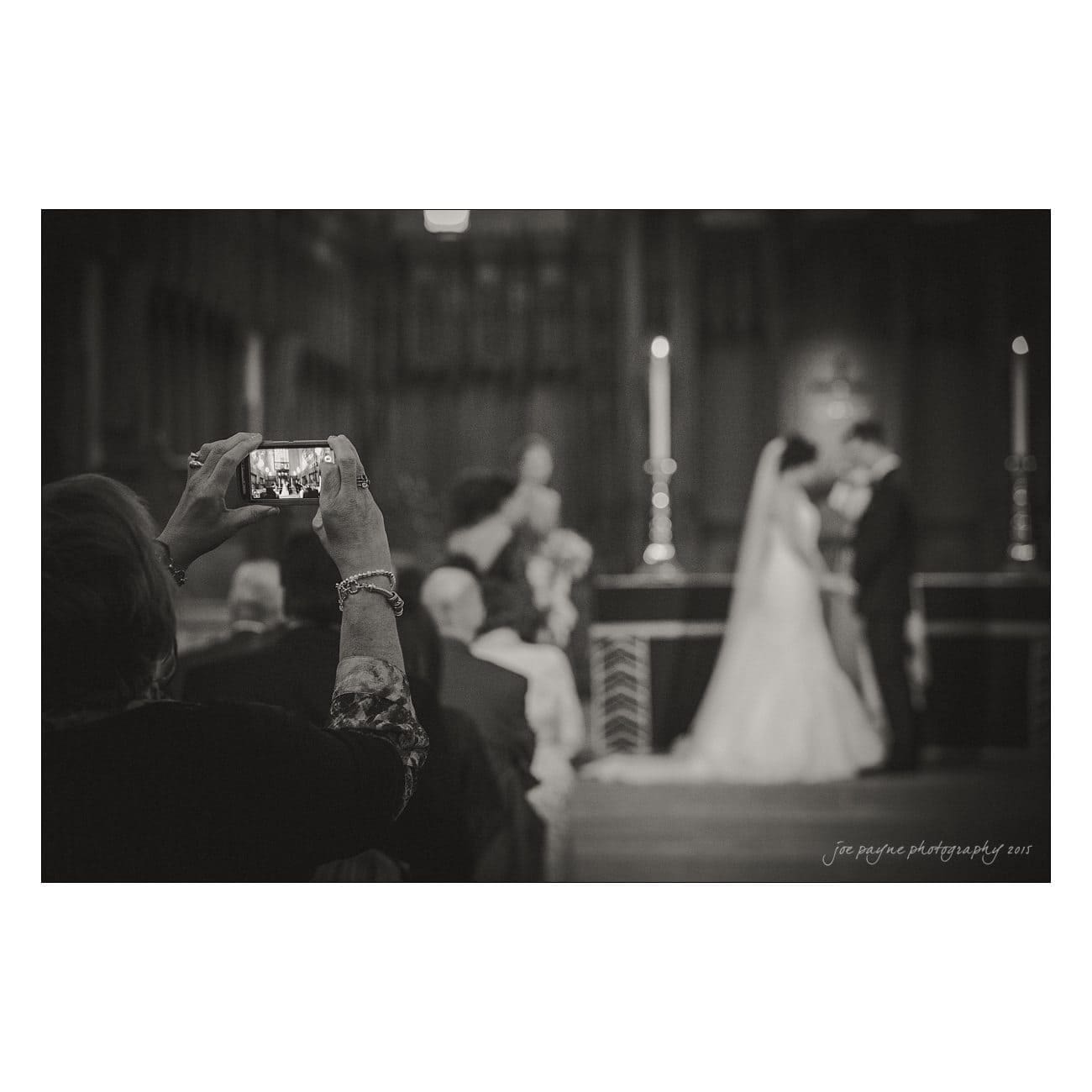 A black and white photo of a couple kissing at their wedding ceremony. A person in the foreground captures the moment on a smartphone. The setting is an indoor venue with dim lighting and candles.