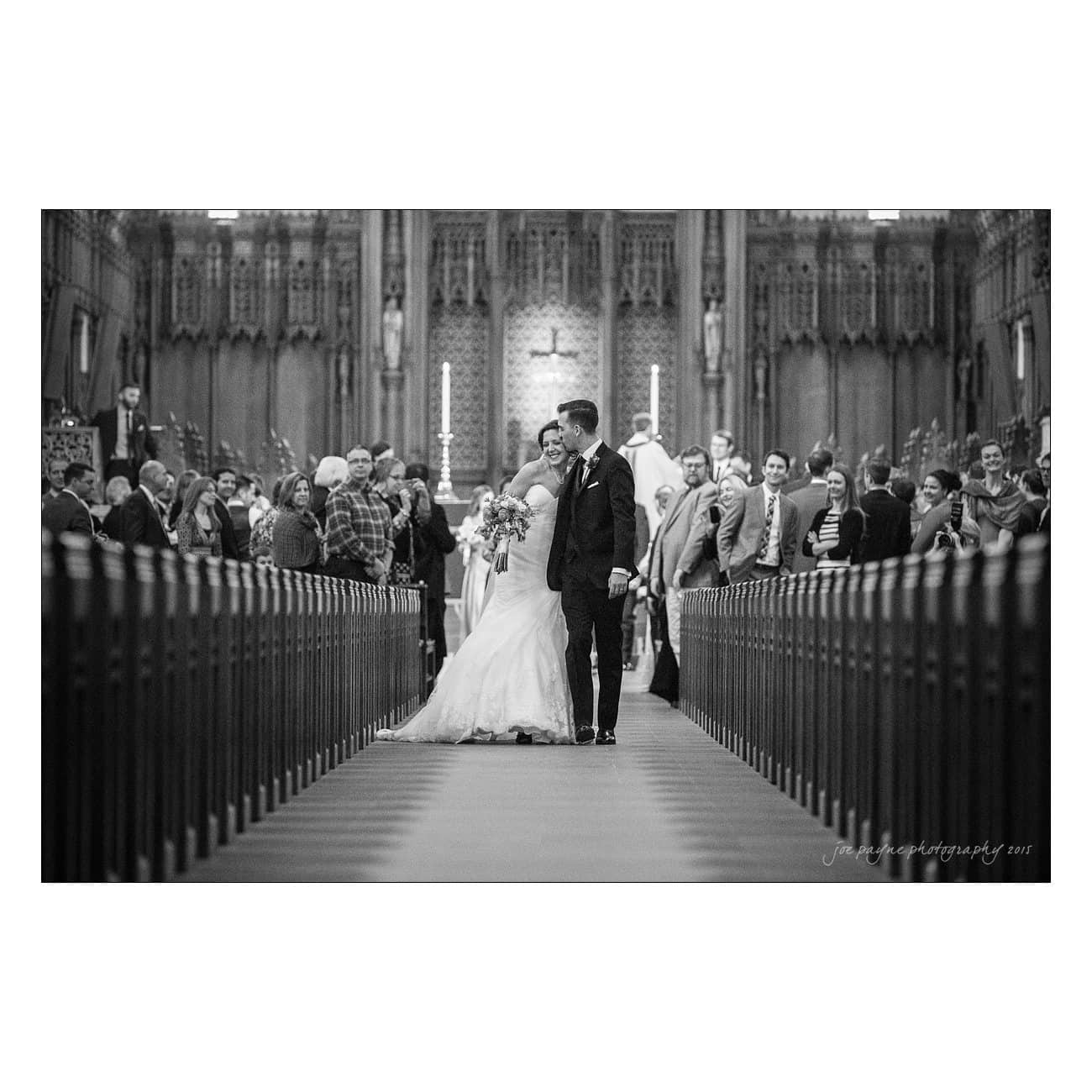 A bride and groom walk arm-in-arm down the aisle of a large, ornate church. They are smiling at each other, surrounded by seated guests who are watching the couple. Candles and intricate architectural details are visible in the background.
