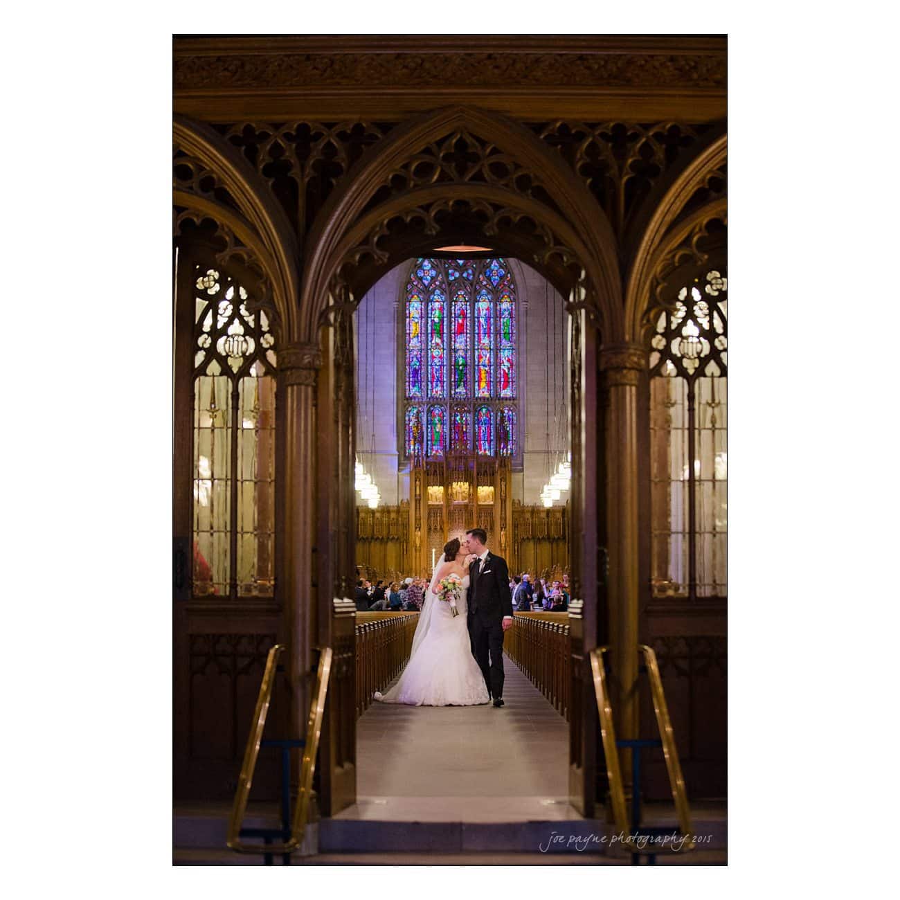 A bride and groom share a kiss at the front of a grand church. They stand under ornate wooden arches, with a stained-glass window behind them casting colorful light into the space. Guests are seated along the aisle.