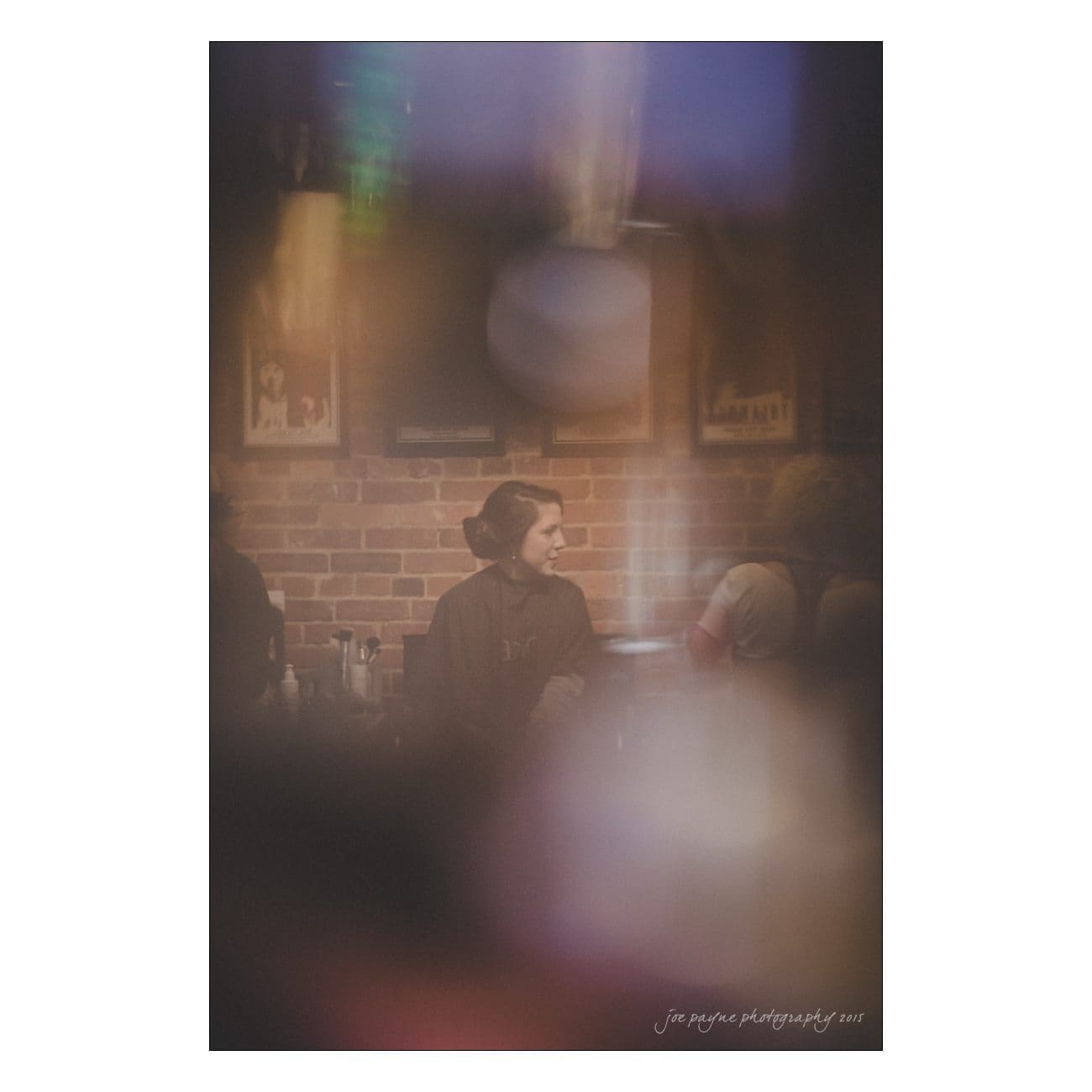A dimly lit, blurred photo of a person with short hair sitting in a cozy brick-walled room. The soft-focus effect creates a dreamy atmosphere with warm tones and hints of framed pictures on the wall.