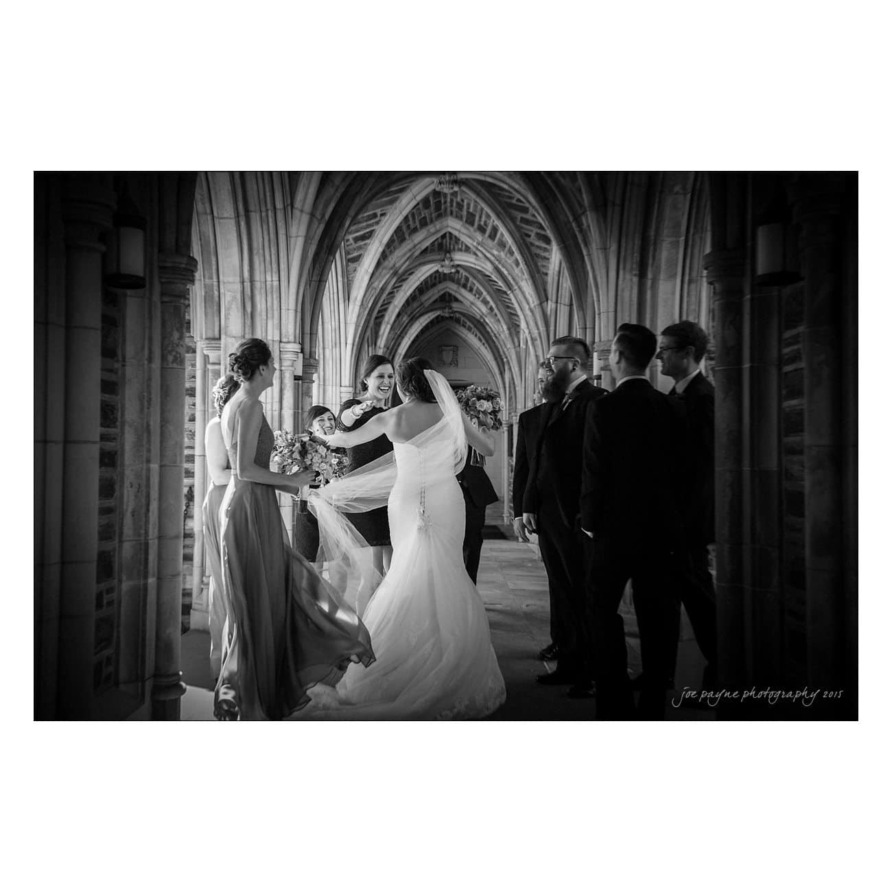 A black and white photo of a bride in a wedding dress hugging a bridesmaid in a long gown, surrounded by the bridal party in a gothic-style arched hallway. The atmosphere is joyful and celebratory.