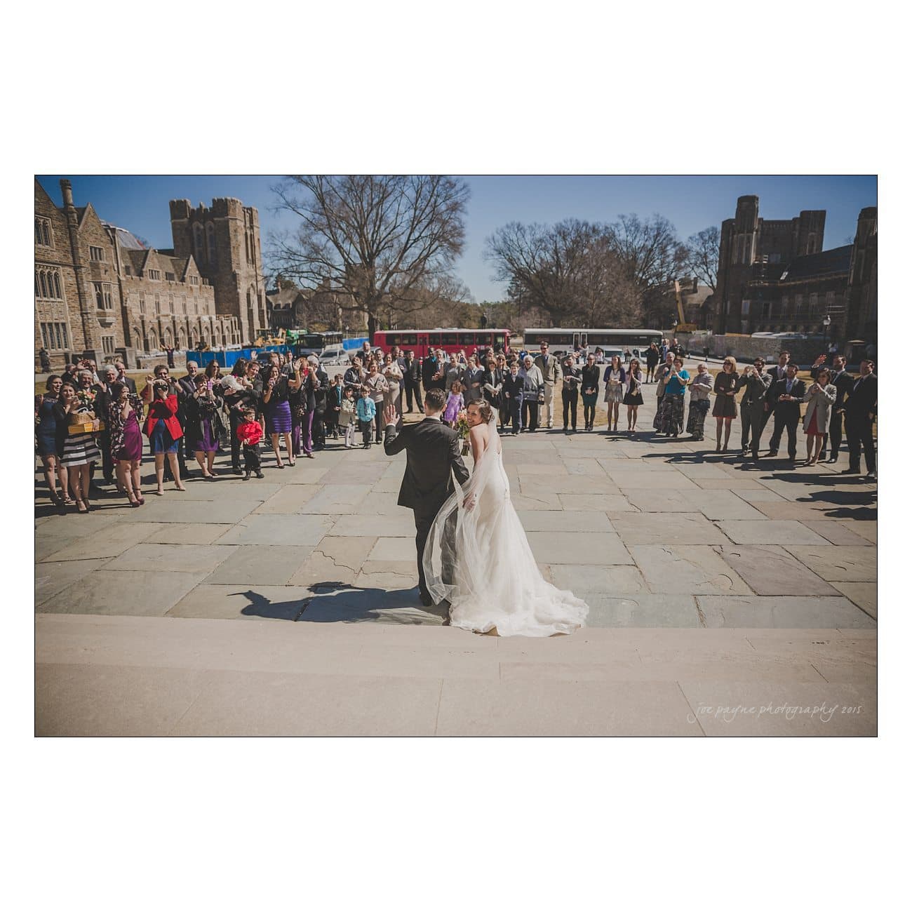 A newlywed couple walks arm in arm across a stone courtyard, surrounded by a large crowd of guests. The scene is outdoors, with a backdrop of historic stone buildings and barren trees under a clear sky.