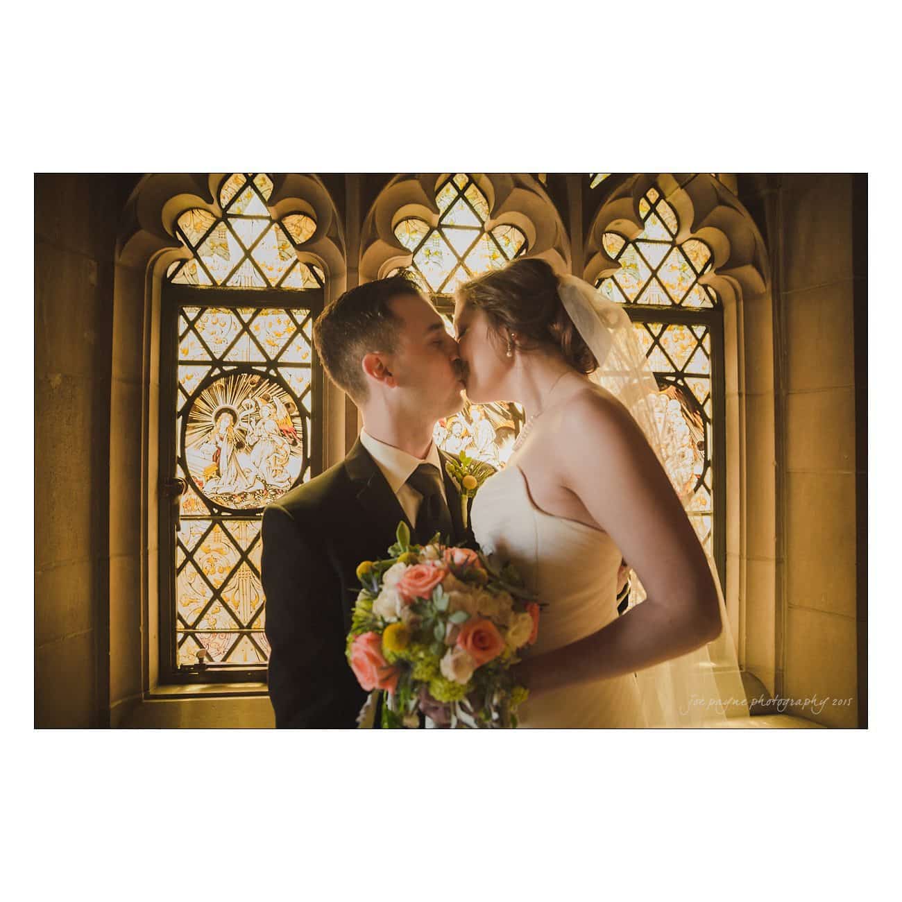 A bride and groom share a kiss in front of an ornate stained glass window. The bride holds a bouquet of pink and white flowers, and both are elegantly dressed for their wedding.