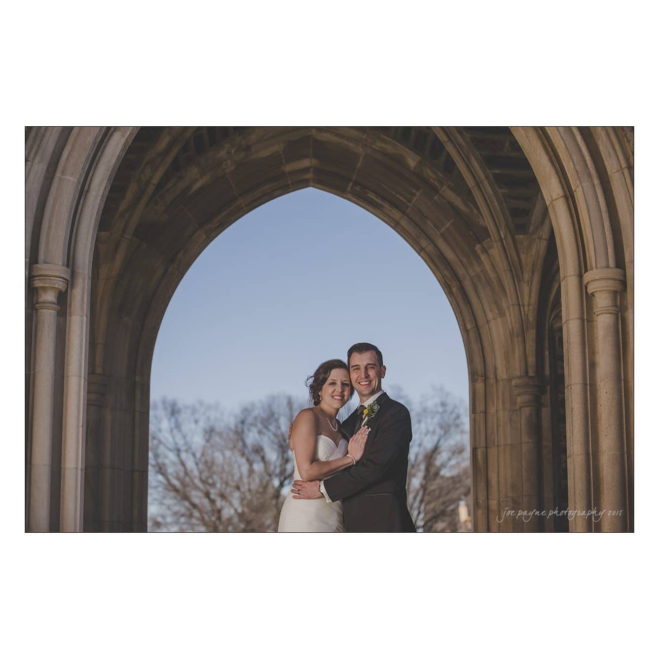 A couple in wedding attire smiles and poses together under a stone archway with a clear sky and trees in the background.
