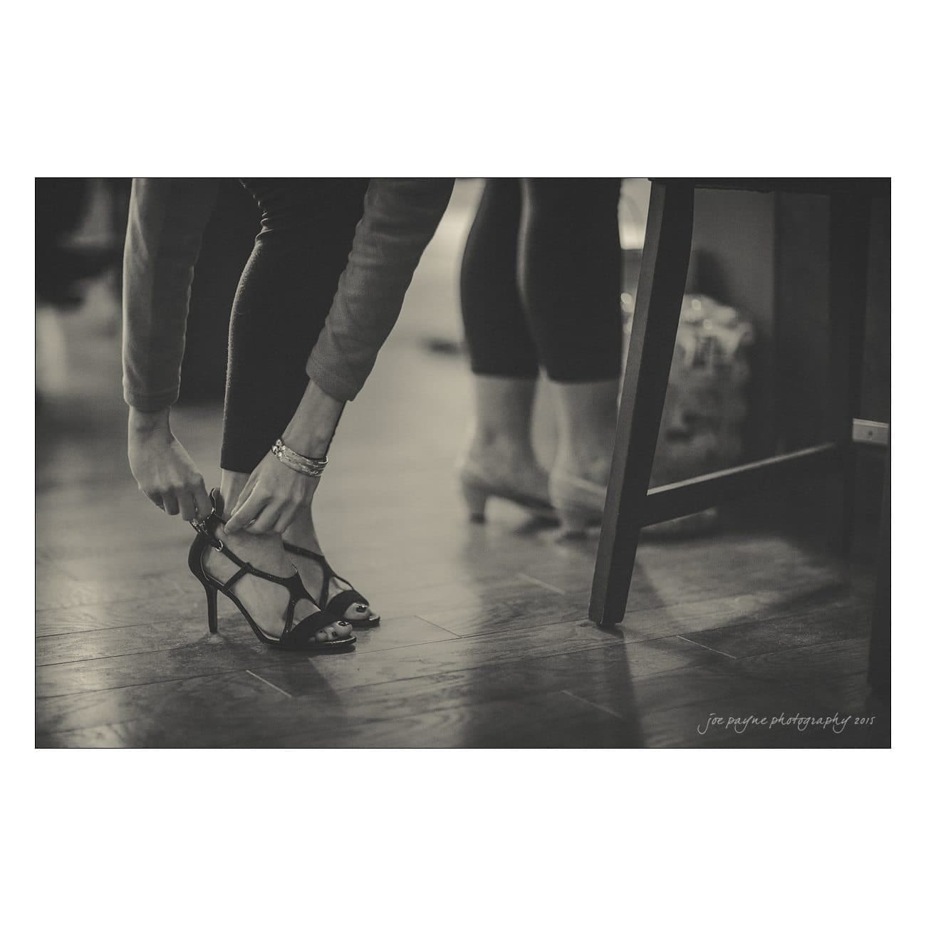 A person in leggings and a long-sleeved top bends down to adjust high-heeled sandals. Their hands hold the ankle straps while another pair of heels is visible in the background. The scene is in black and white.