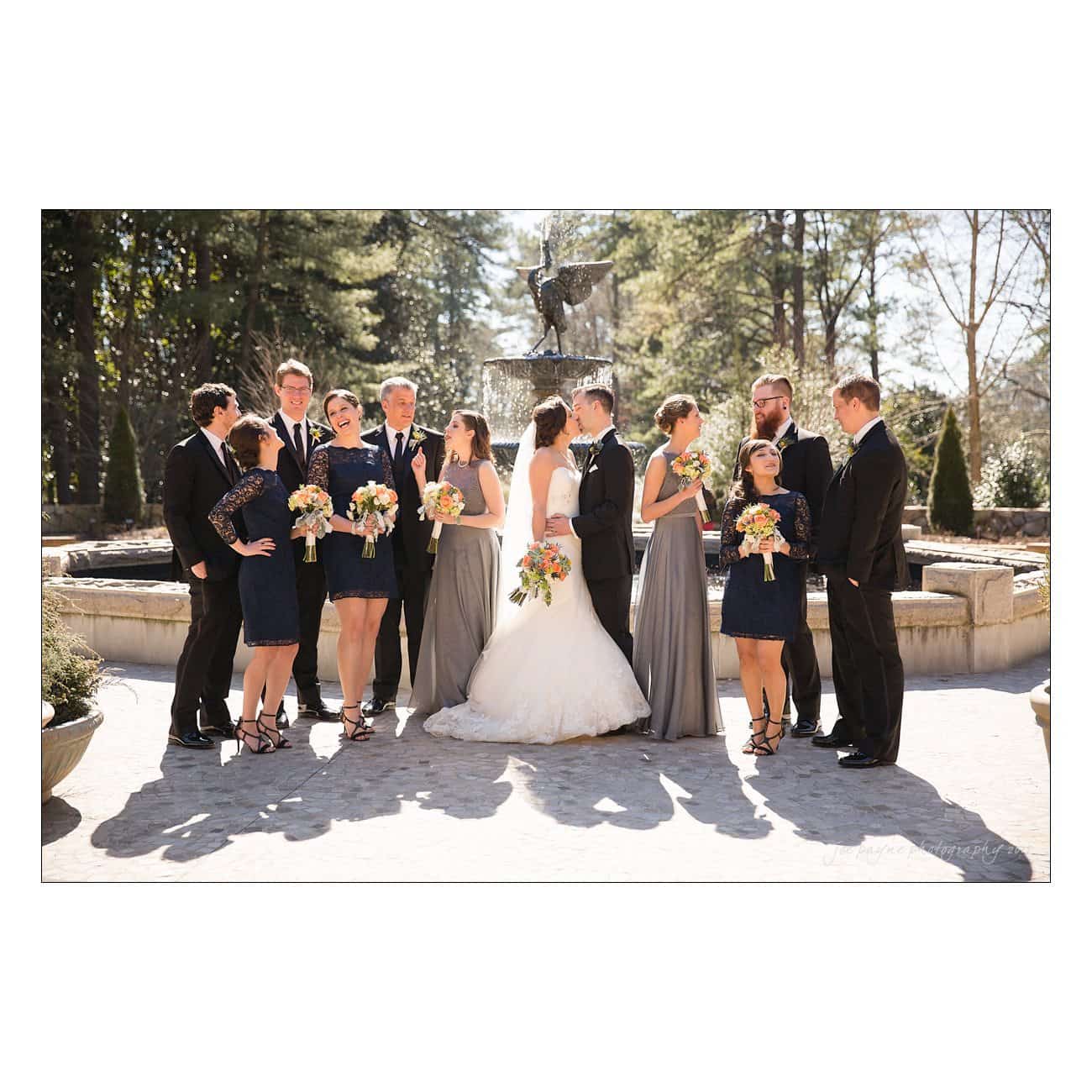 A wedding party poses outdoors near a fountain. The group includes the bride in a white dress, the groom in a black suit, bridesmaids in gray dresses, and groomsmen in dark suits. Theyre all smiling and holding bouquets.