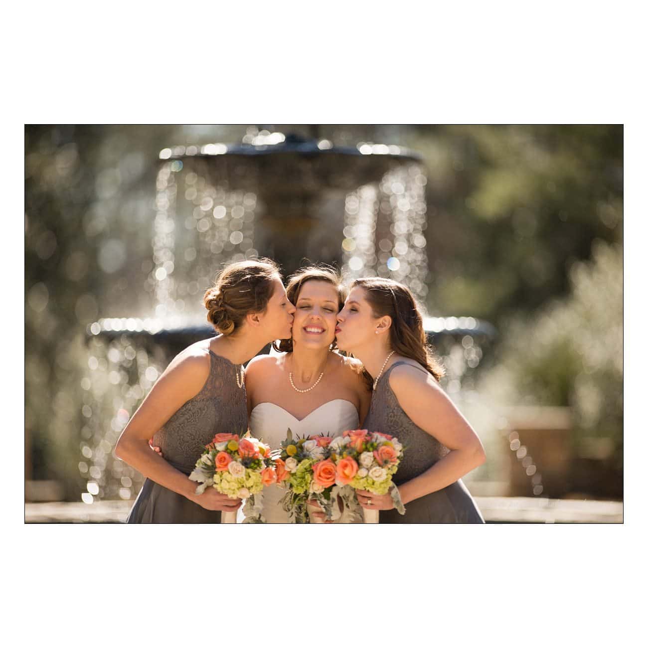 A bride in a white dress, holding a bouquet of flowers, is flanked by two bridesmaids in grey dresses. They are happily kissing her cheeks. A blurred fountain is in the background under sunny, clear skies.