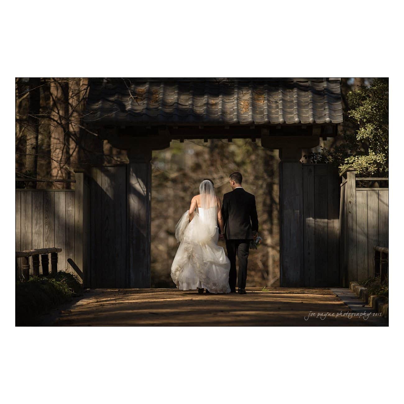 A bride and groom walk hand in hand under a rustic wooden gate. The bride wears a white gown and veil, and the groom wears a black suit. Sunlight filters through the trees, creating a warm, romantic atmosphere.