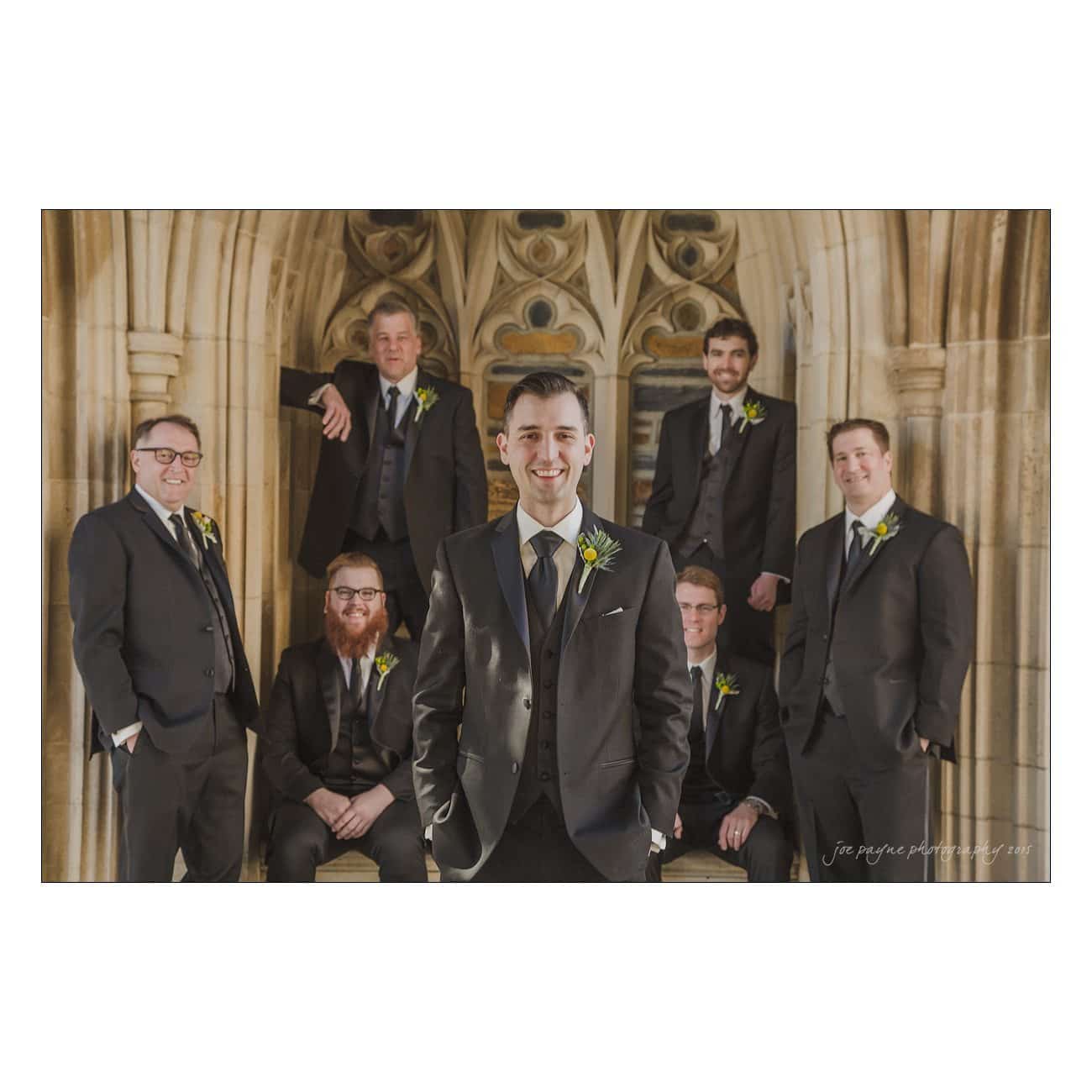 A group of seven men in suits pose together in an ornate stone archway. The man in the foreground stands smiling, while the others are arranged behind him, some seated and others standing, all wearing boutonnieres.