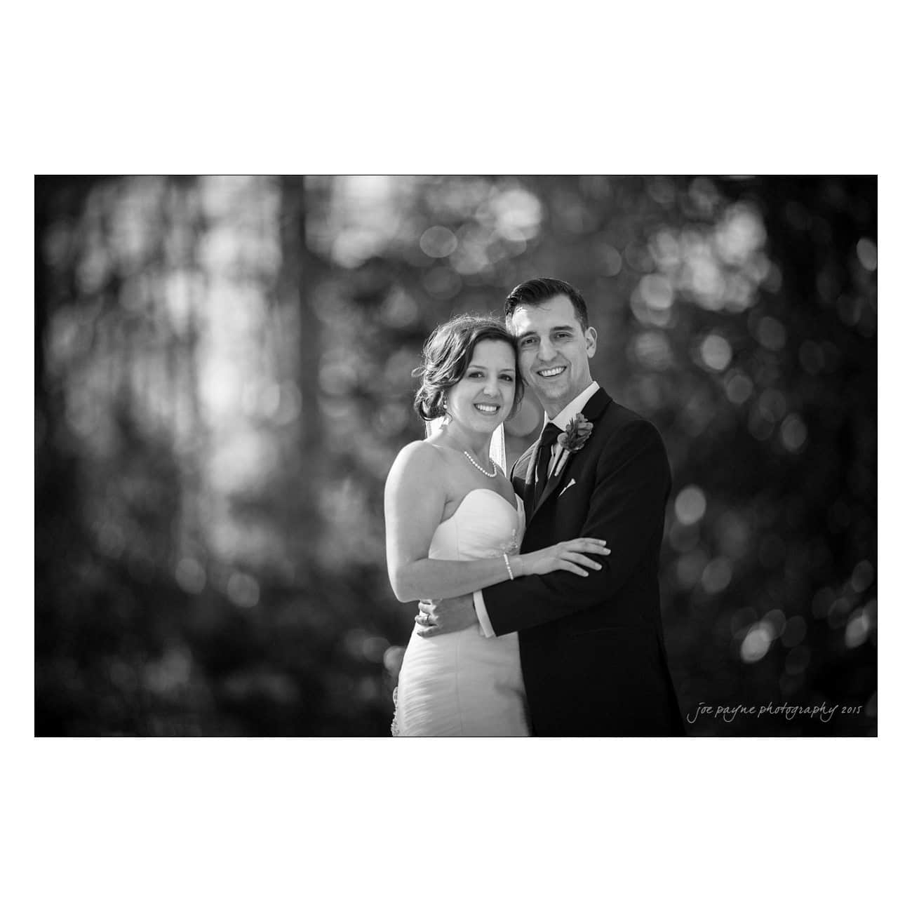 A smiling couple embraces in a black and white portrait. The background is softly blurred, highlighting their expressions. Both are dressed elegantly, suggesting a formal occasion.