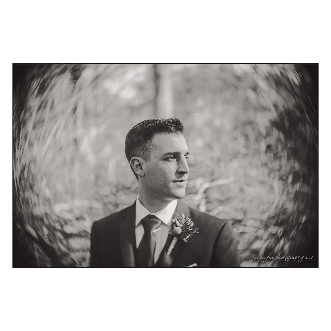 Black and white portrait of a man in a suit looking to the side. He wears a boutonnière on his lapel. The background is blurred with a circular motion effect.
