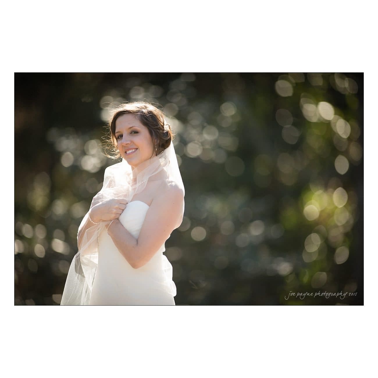 A bride in a white wedding dress and veil smiles softly while standing outdoors. The background is blurred with circular light bokeh, creating a dreamy effect.