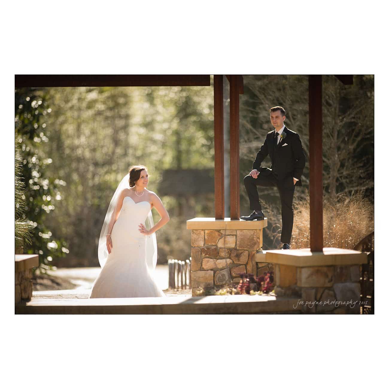 A bride in a white gown stands with hands on hips, facing a groom in a dark suit who poses with a foot on a stone ledge. They are outdoors under a wooden structure, surrounded by sunlit greenery.