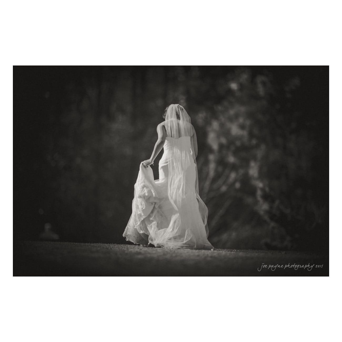A bride in a flowing wedding dress walks outdoors, away from the camera. She holds up the hem of her dress with one hand. The background is a serene, softly blurred forest setting.