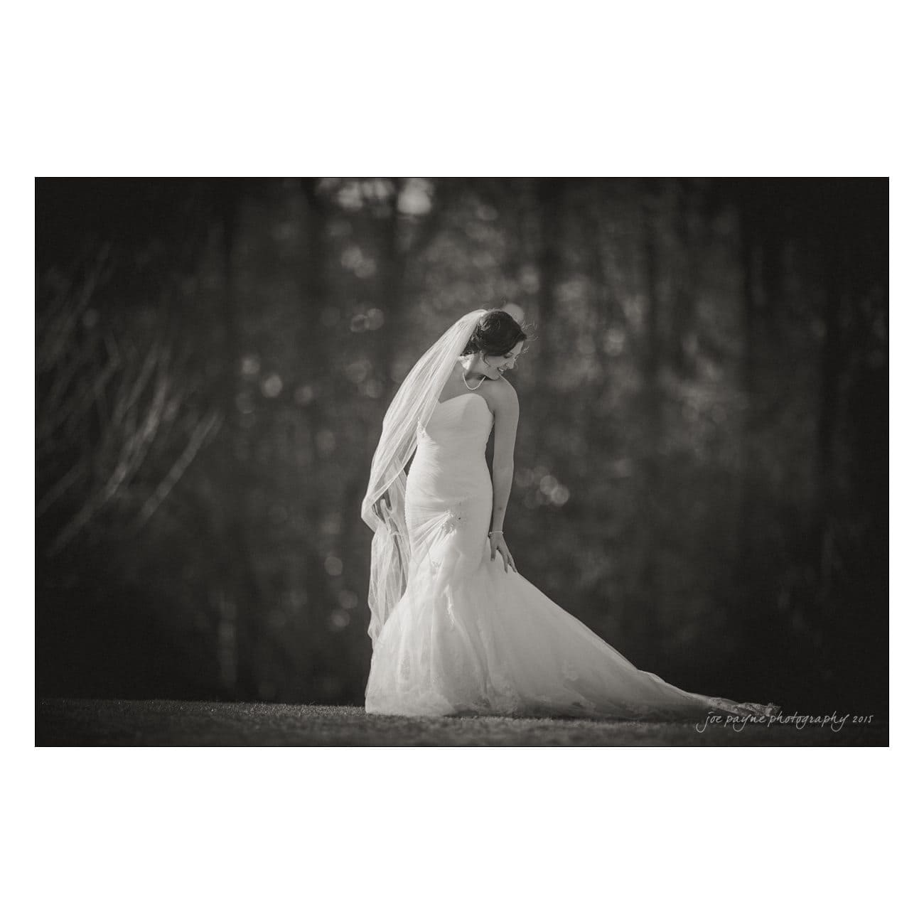 Black and white photo of a bride in a long, elegant wedding gown with a veil, standing outdoors. She is looking over her shoulder in a serene forest setting.