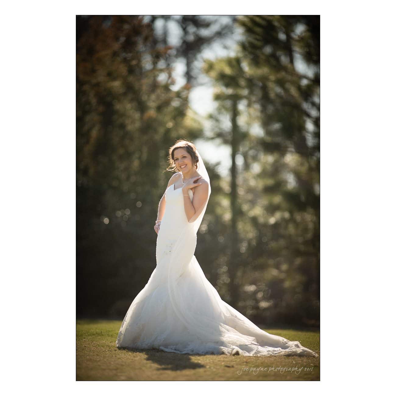 A bride in a white wedding dress poses outdoors on a sunny day. She stands on grass with trees in the background, looking towards the camera and smiling.
