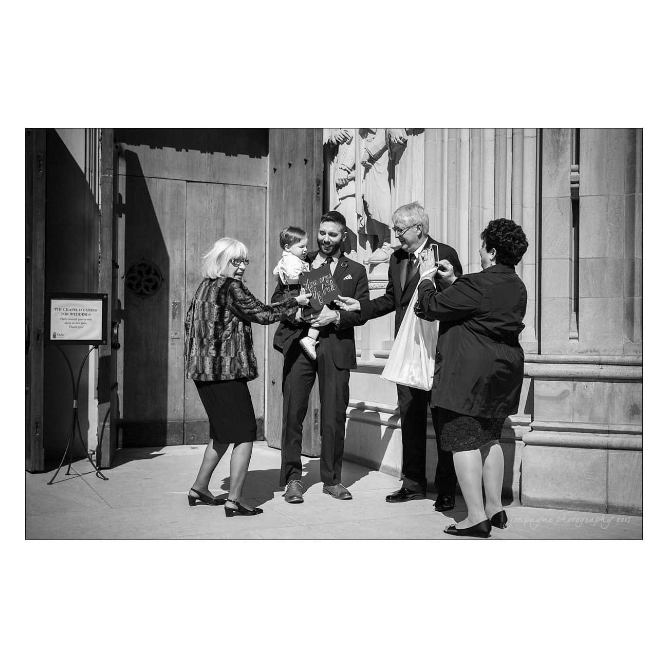 A black and white photo of a joyous family moment outside a building. Five people are gathered, including a child held by a smiling man. Two women and an older man are also present, sharing in the happy occasion under the sunlight.