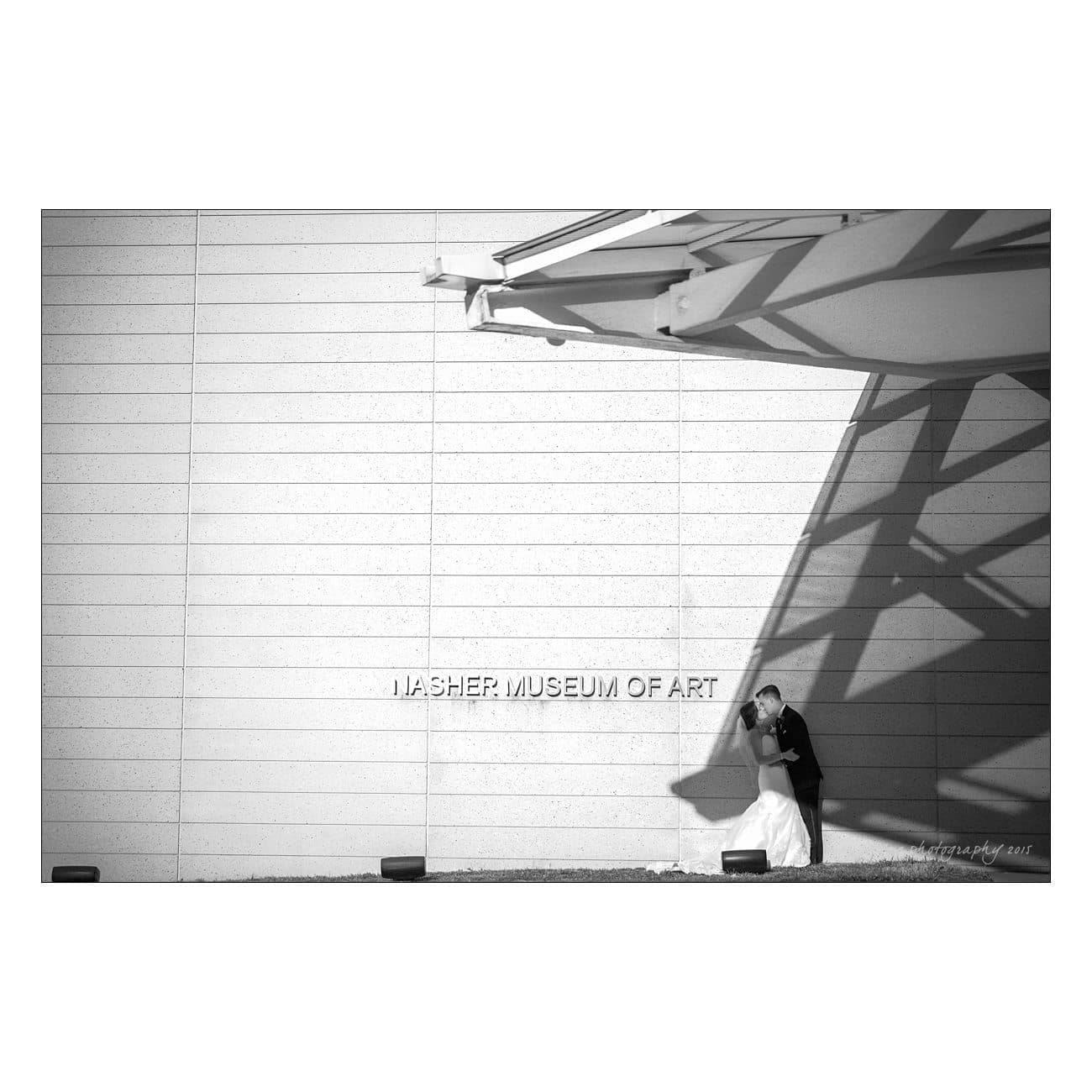 Black and white photo of a couple embracing against a wall at the Nasher Museum of Art. Bold shadows of architectural elements fall across the scene, creating a dramatic effect.