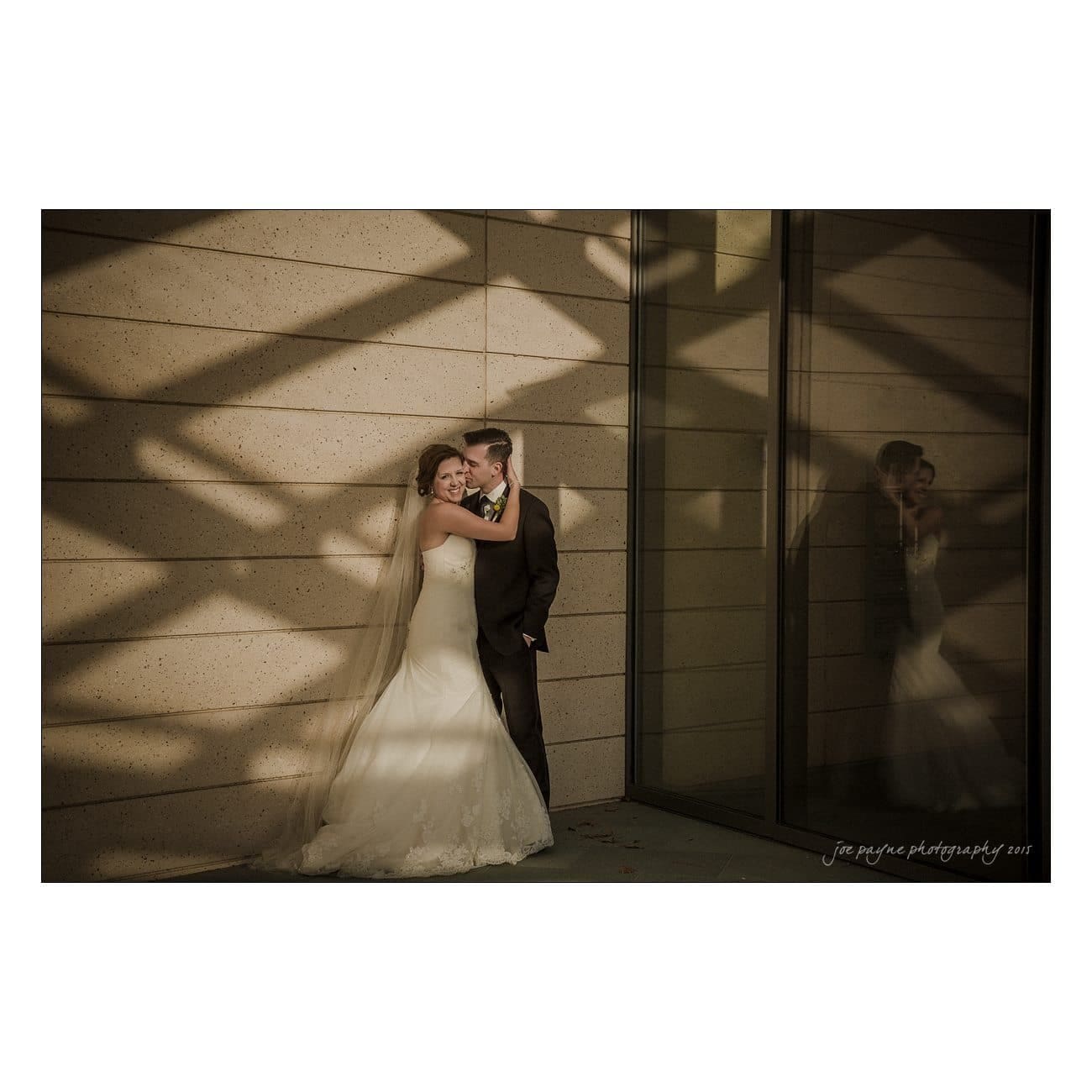 A bride and groom stand close together in a corner, smiling. The bride wears a long wedding dress with a veil, and the groom is in a suit. Shadows from a lattice create patterns on the wall and floor. Their reflection is visible in a glass panel.