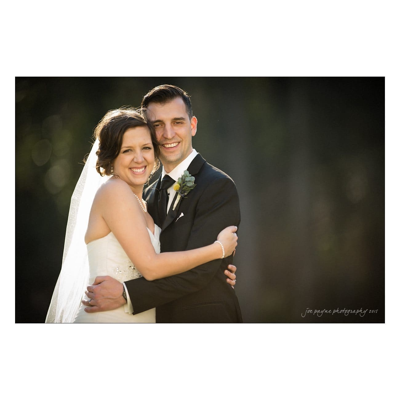 A bride and groom smiling and embracing each other outdoors. The bride is wearing a white dress with a veil, and the groom is in a black suit with a boutonniere. The background is softly blurred greenery.