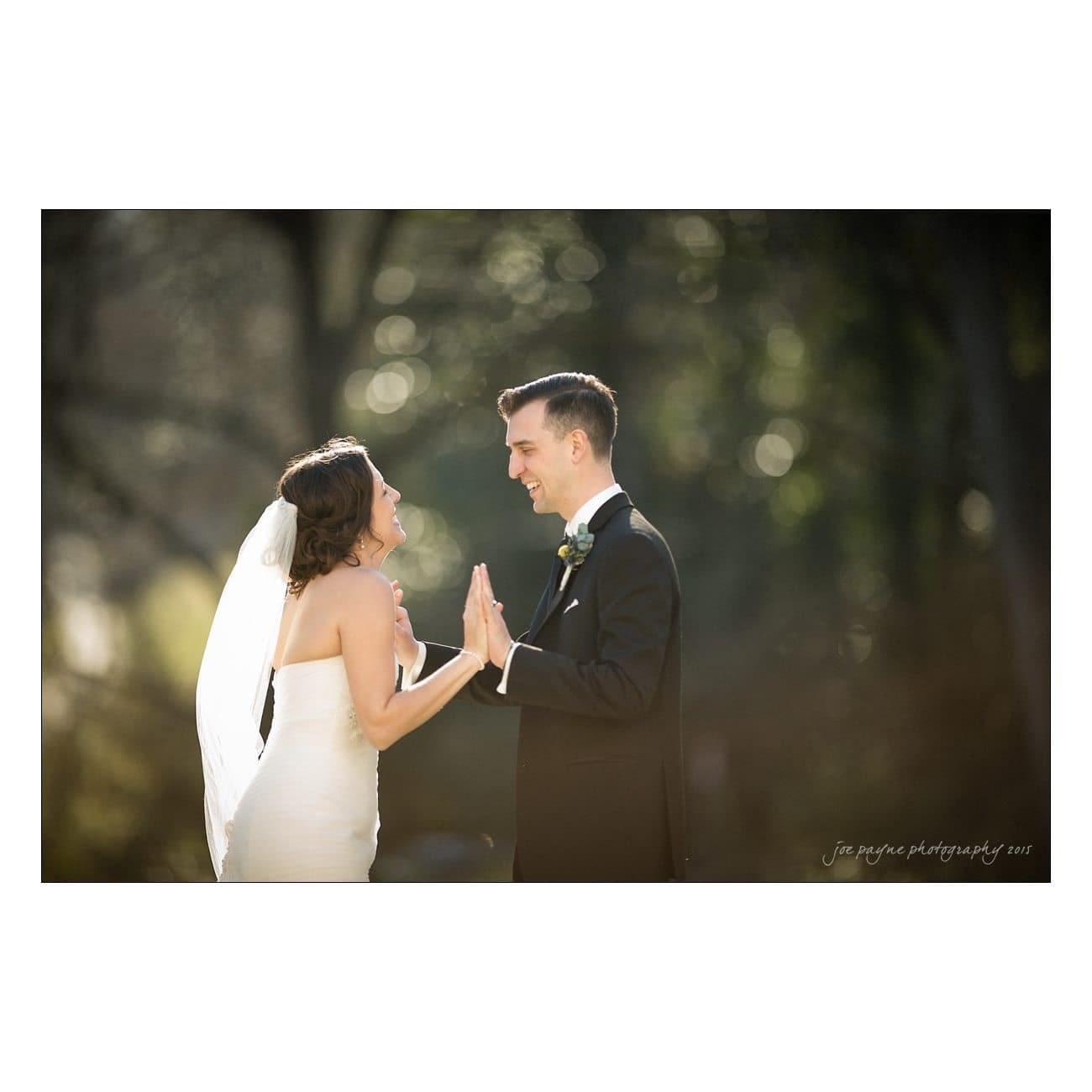 A bride and groom are standing outdoors, facing each other with hands touching. The bride is wearing a white strapless dress and veil, and the groom is in a black suit. The background is softly blurred trees, creating a warm, joyful atmosphere.