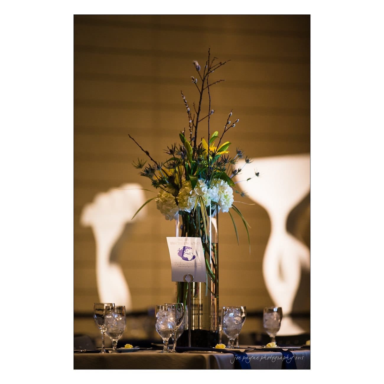 A tall floral centerpiece with white hydrangeas, greenery, and twigs in a glass vase on a table. Water glasses surround the arrangement, and a card with the letter E is attached. Shadows cast on the wall in the background.