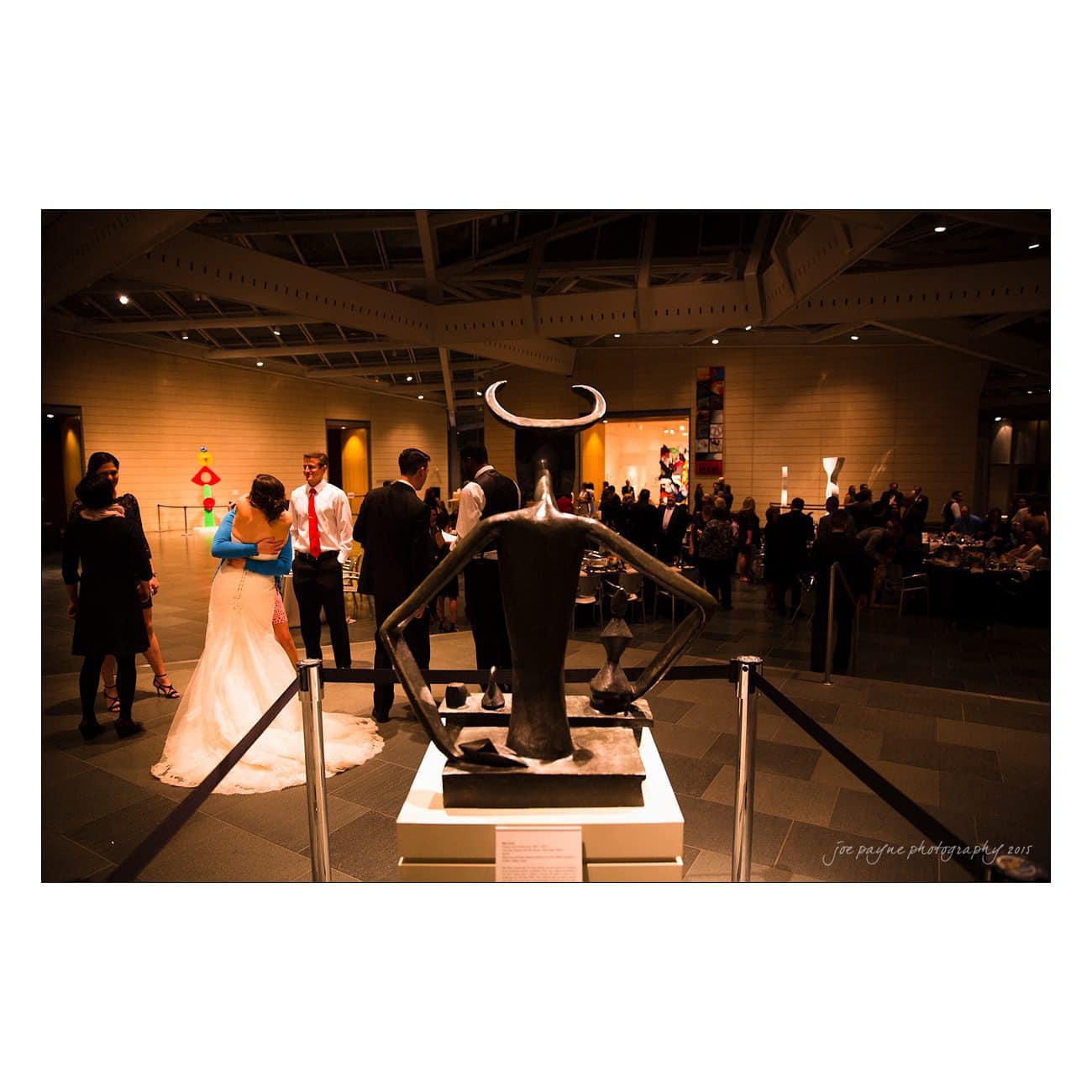 A formal event in a gallery setting, with guests socializing in elegant attire. The foreground features a modern sculpture, and colorful abstract art pieces adorn the background walls. The lighting is warm and atmospheric.