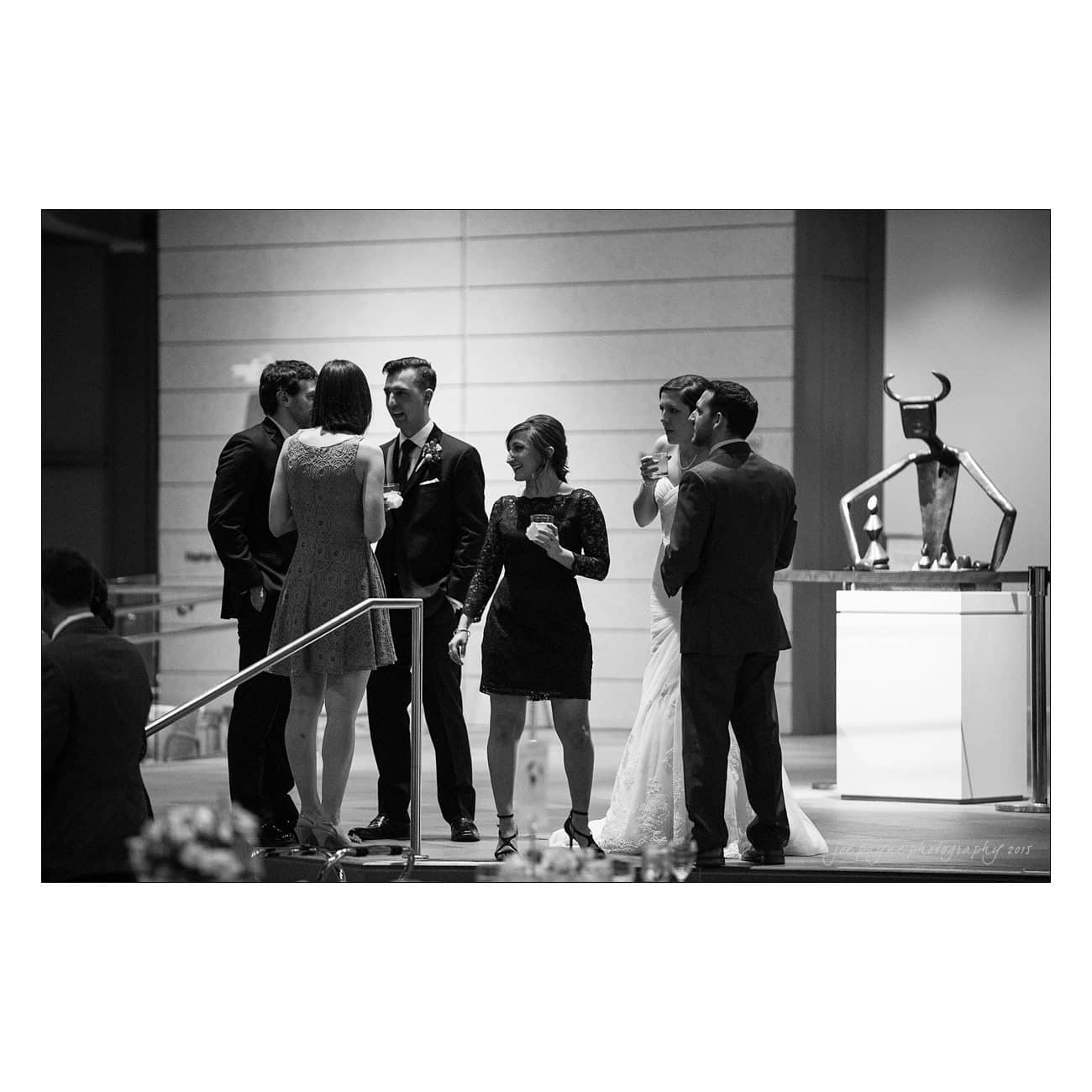 A group of people in formal attire gather on a stage, engaged in conversation. The setting appears to be a modern, indoor venue. A statue with bull-like features is visible on a pedestal nearby. The image is in black and white.