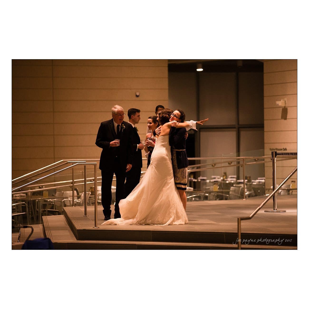 A bride in a white gown stands on a small stage with several people dressed in formal attire. One person points off to the side while others hold drinks. The setting appears to be indoors with metal chairs and tables in the background.