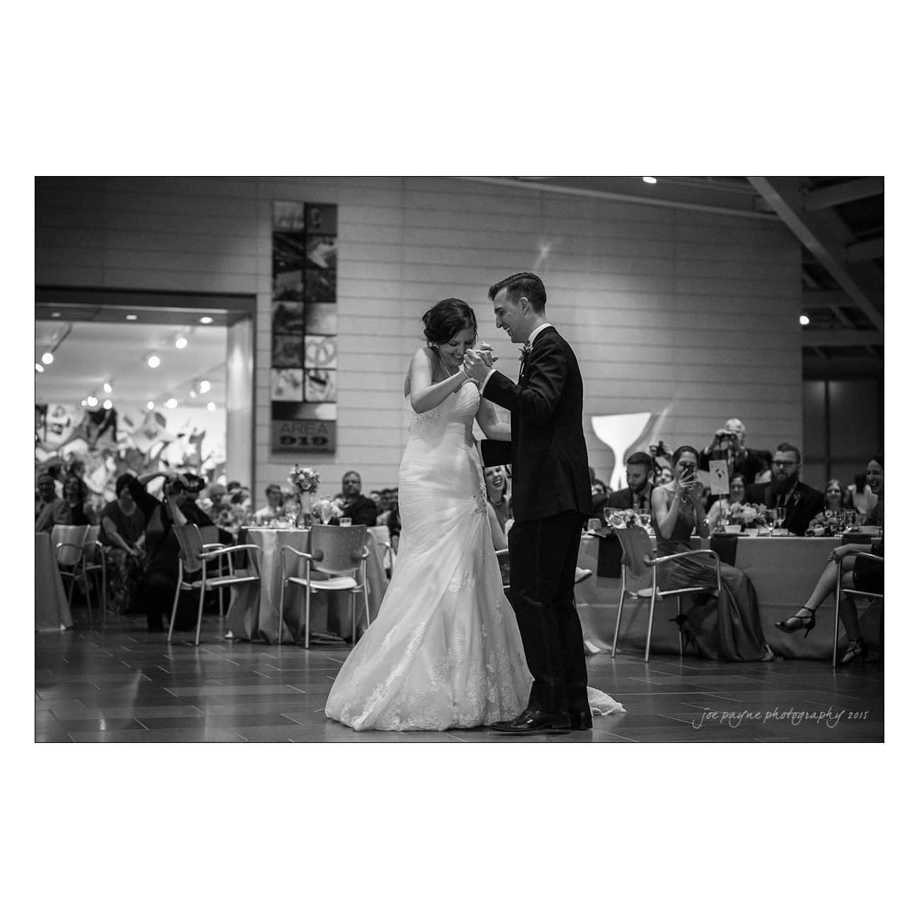 A bride and groom share a dance in a spacious reception hall. The bride wears a long, white gown, and the groom is in a suit. Guests seated at round tables watch and take pictures. The atmosphere is joyful and celebratory.