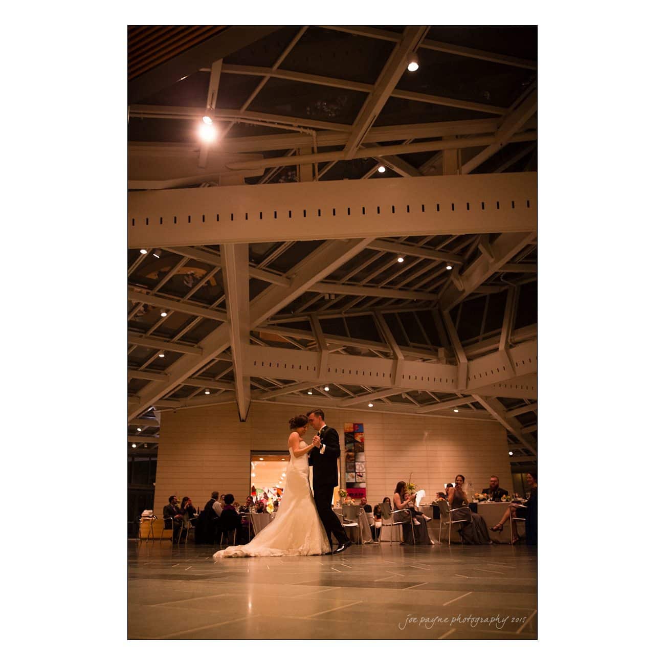 A bride in a white gown and a groom in a dark suit share their first dance in a modern, architecturally interesting hall with high ceilings. Seated guests watch the couple from tables around the room.