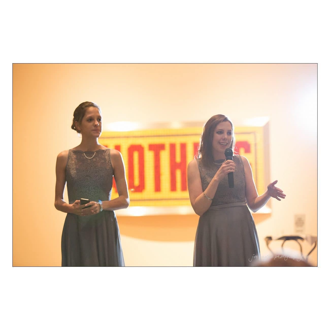 Two women in grey dresses stand and speak into a microphone in a softly lit room. A brightly lit sign is in the background.