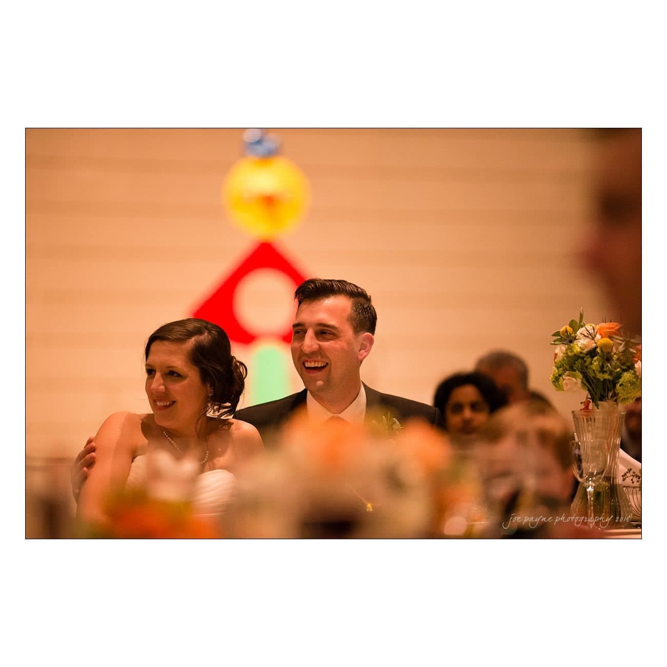 A smiling couple sits together at an indoor event, surrounded by a warm atmosphere. A colorful abstract artwork hangs in the background, and a vase of flowers is visible nearby.