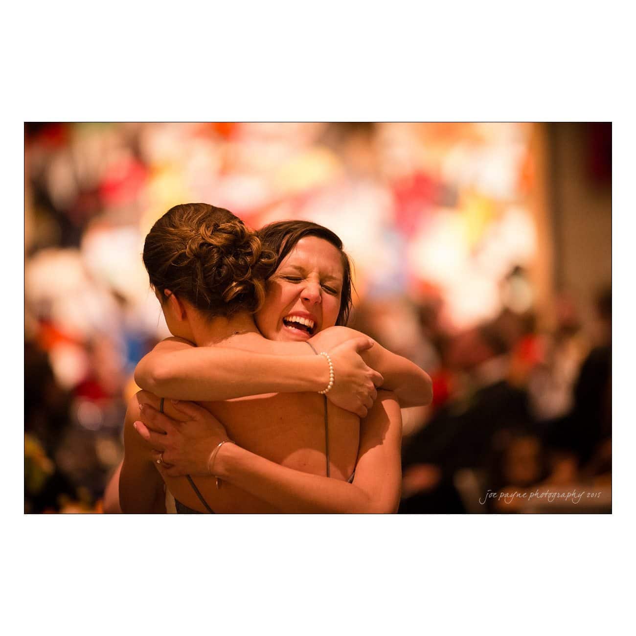 Two women are hugging closely in a warmly lit setting. One woman is smiling broadly with her eyes closed, expressing happiness or excitement. The background is blurred with colorful lights, suggesting a celebratory atmosphere.