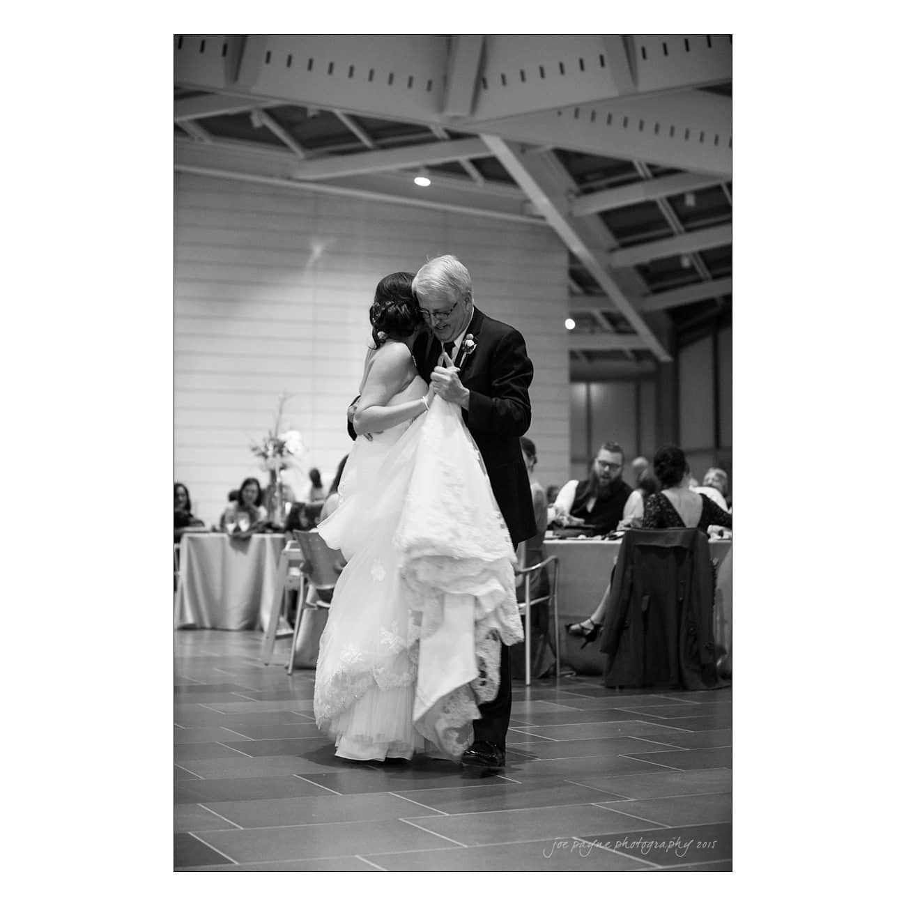 A black and white photo of a bride and groom dancing closely at a wedding reception. The bride holds her gown with one hand. Guests seated at tables are visible in the softly lit background.