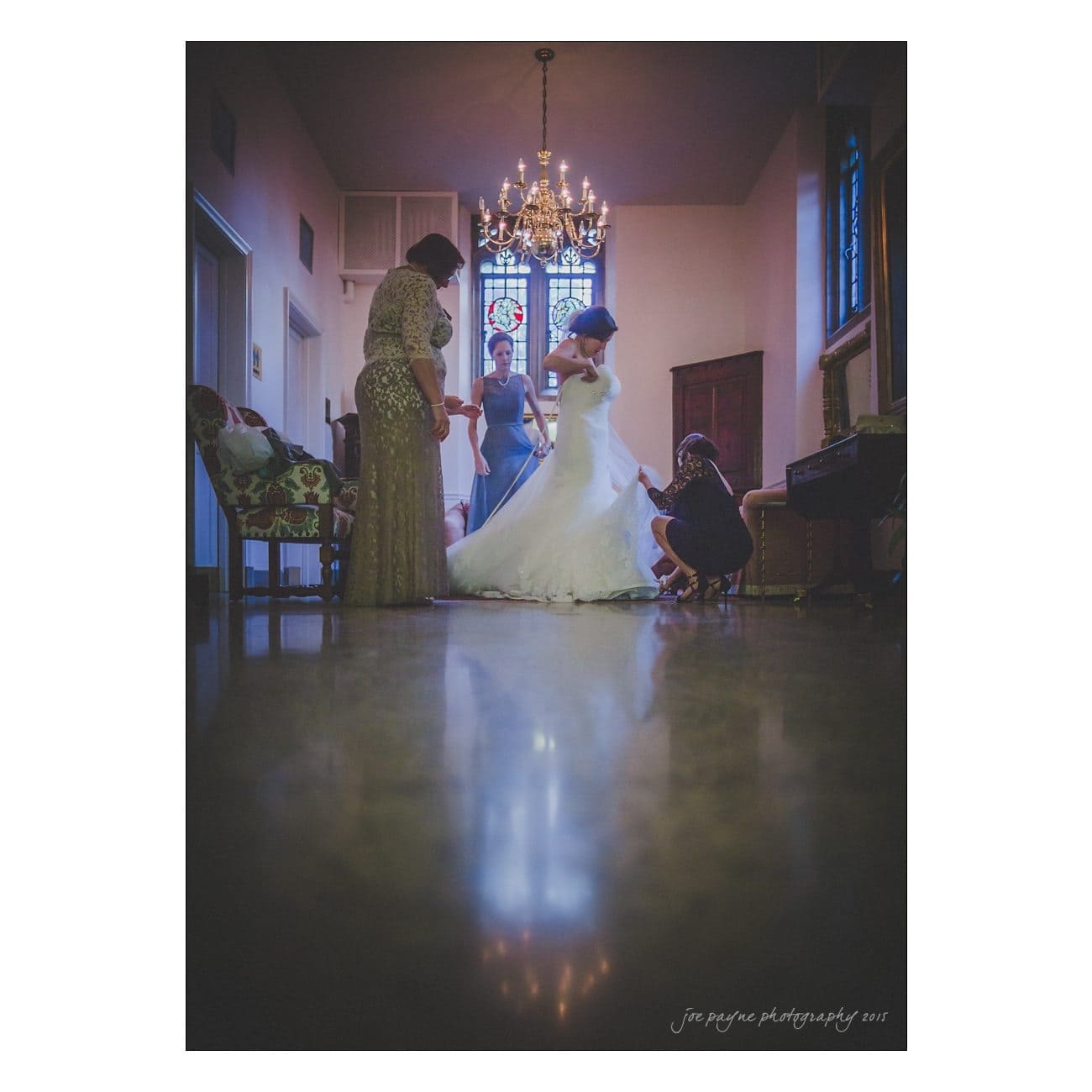 A bride stands in a white wedding gown as three women assist her. The scene is set in a dimly lit room with a chandelier and a stained glass window in the background. The floor reflects the figures, adding depth to the atmosphere.