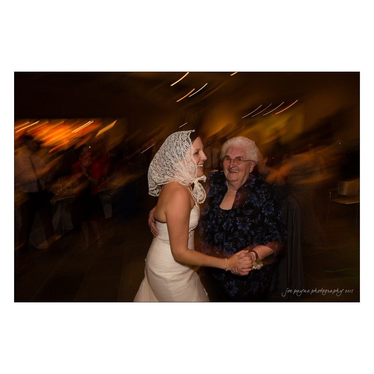 A bride in a white dress and lace headscarf smiles as she dances with an elderly woman wearing a dark dress and glasses. They hold hands and dance in a warmly lit room, surrounded by a blurred background.
