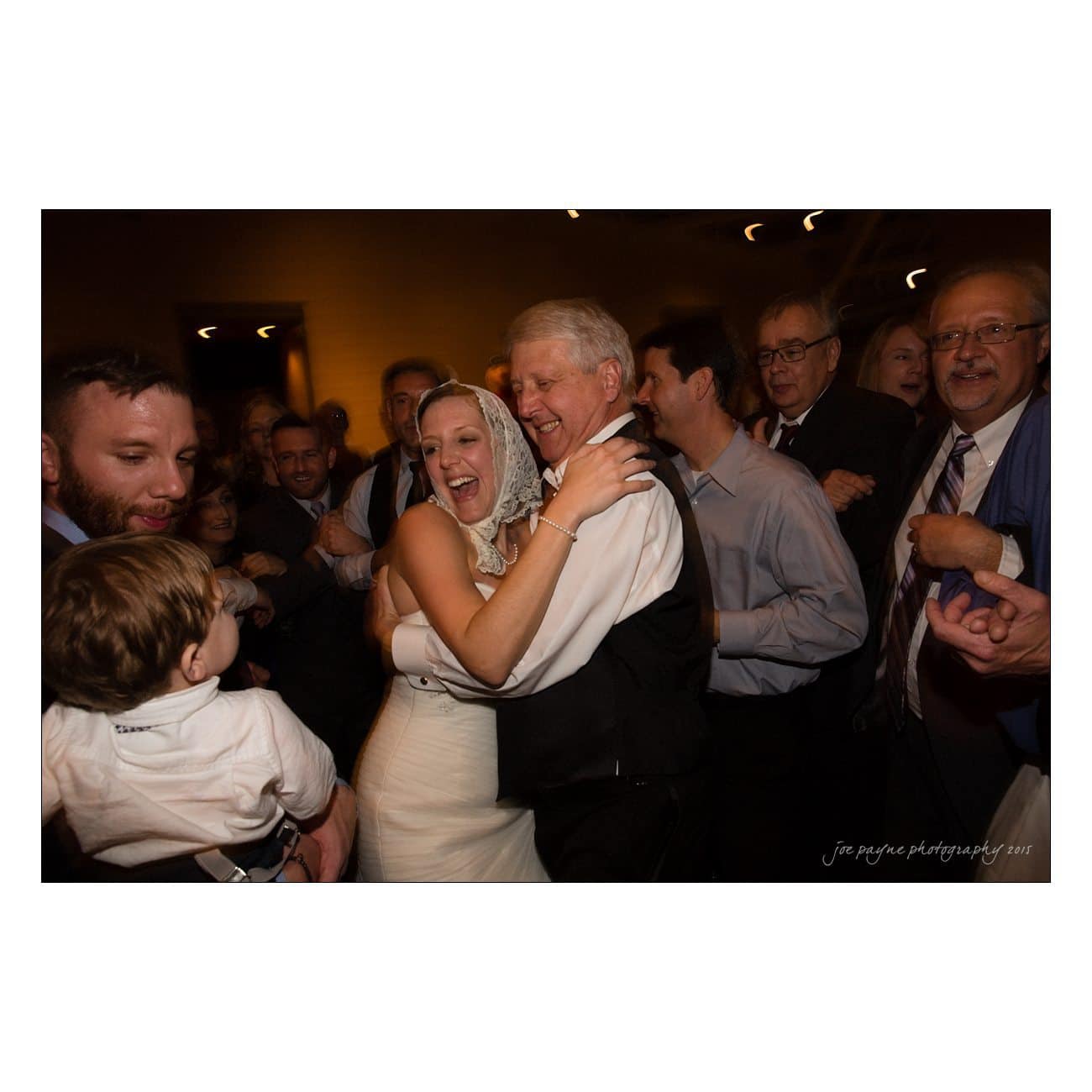 A bride in a white dress and veil joyfully embraces an older man on the dance floor, surrounded by a lively, cheering crowd. A small child is in the foreground. The scene is filled with happiness and celebration.