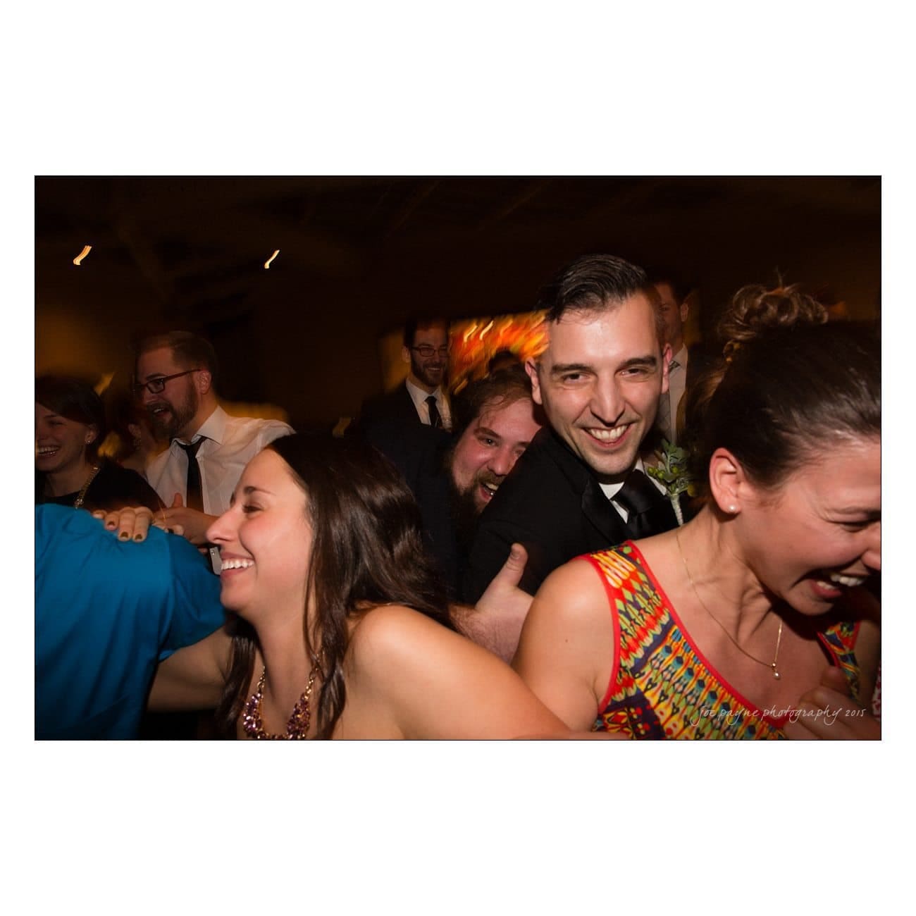 A group of people joyfully dancing in a close line at a celebration. They are smiling and laughing, wearing formal attire. The background is dimly lit, creating a lively and festive atmosphere.