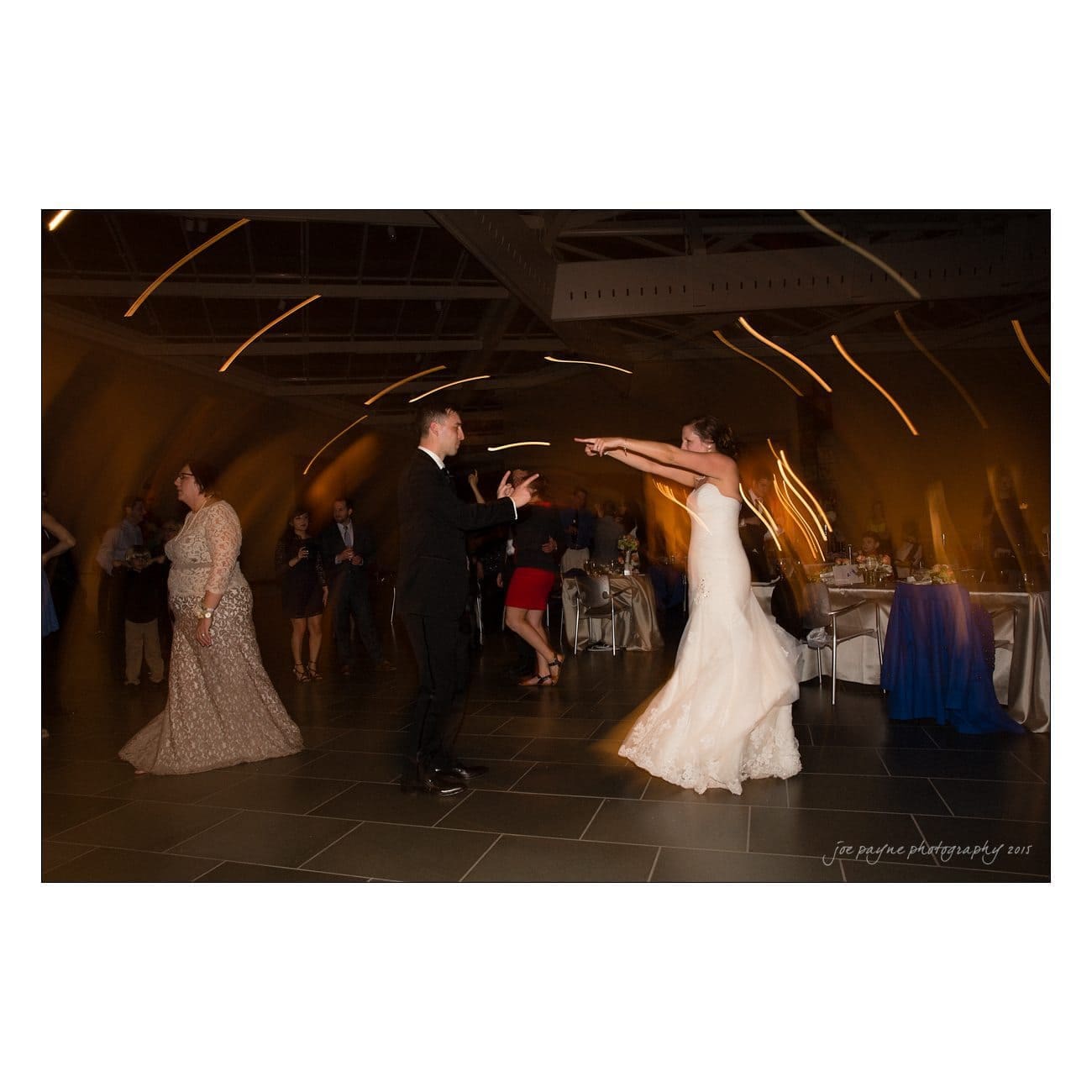 A bride in a wedding gown dances energetically with a man in a suit in a large, dimly lit ballroom. Guests in formal attire are visible in the background. Motion blur adds a dynamic feel to the scene.