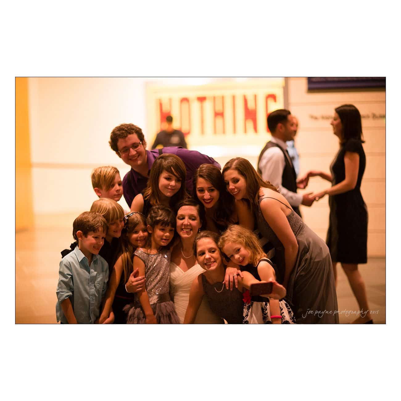 A group of people, including adults and children, pose together for a photo at an indoor event. Some are dressed in formal attire. The background shows a sign with the word NOTHING partially visible.