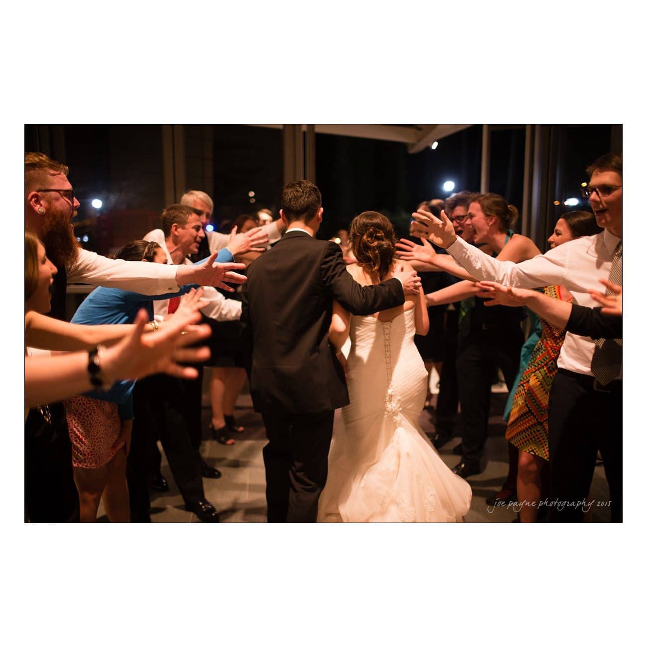A bride and groom walk arm in arm through a joyful crowd of friends and family with arms raised. The bride wears a long white gown, and the groom is in a dark suit. The atmosphere is celebratory and festive.