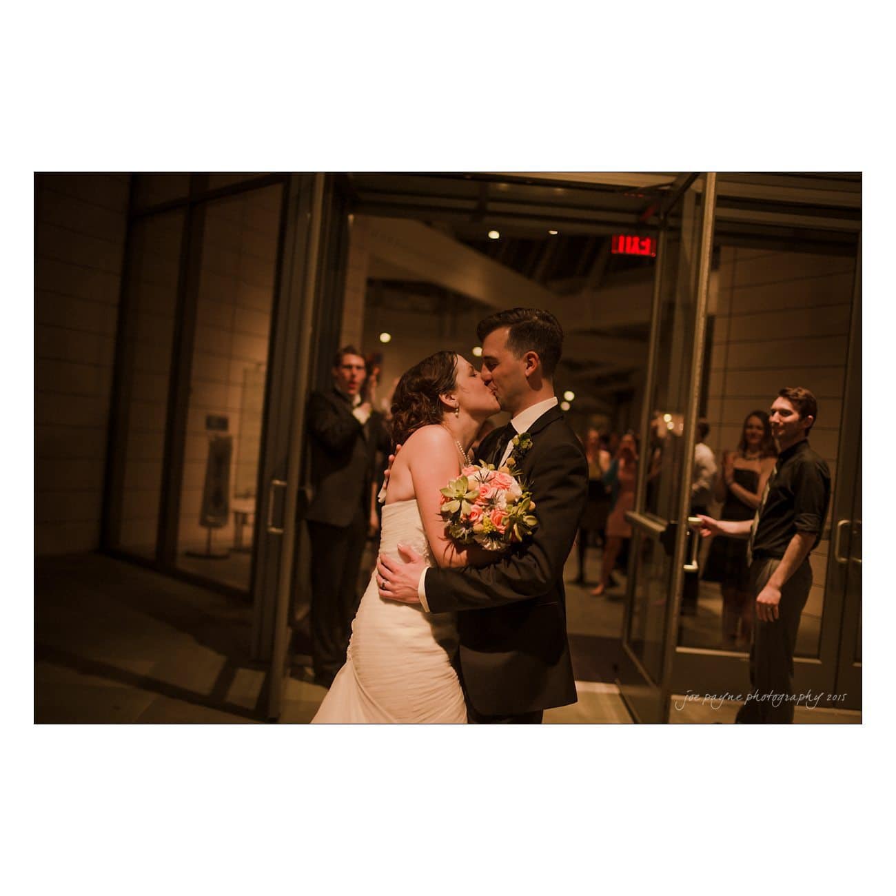 A bride and groom share a kiss at the entrance of a building. The groom holds a bouquet of flowers. People stand in the background, and the scene is warmly lit.
