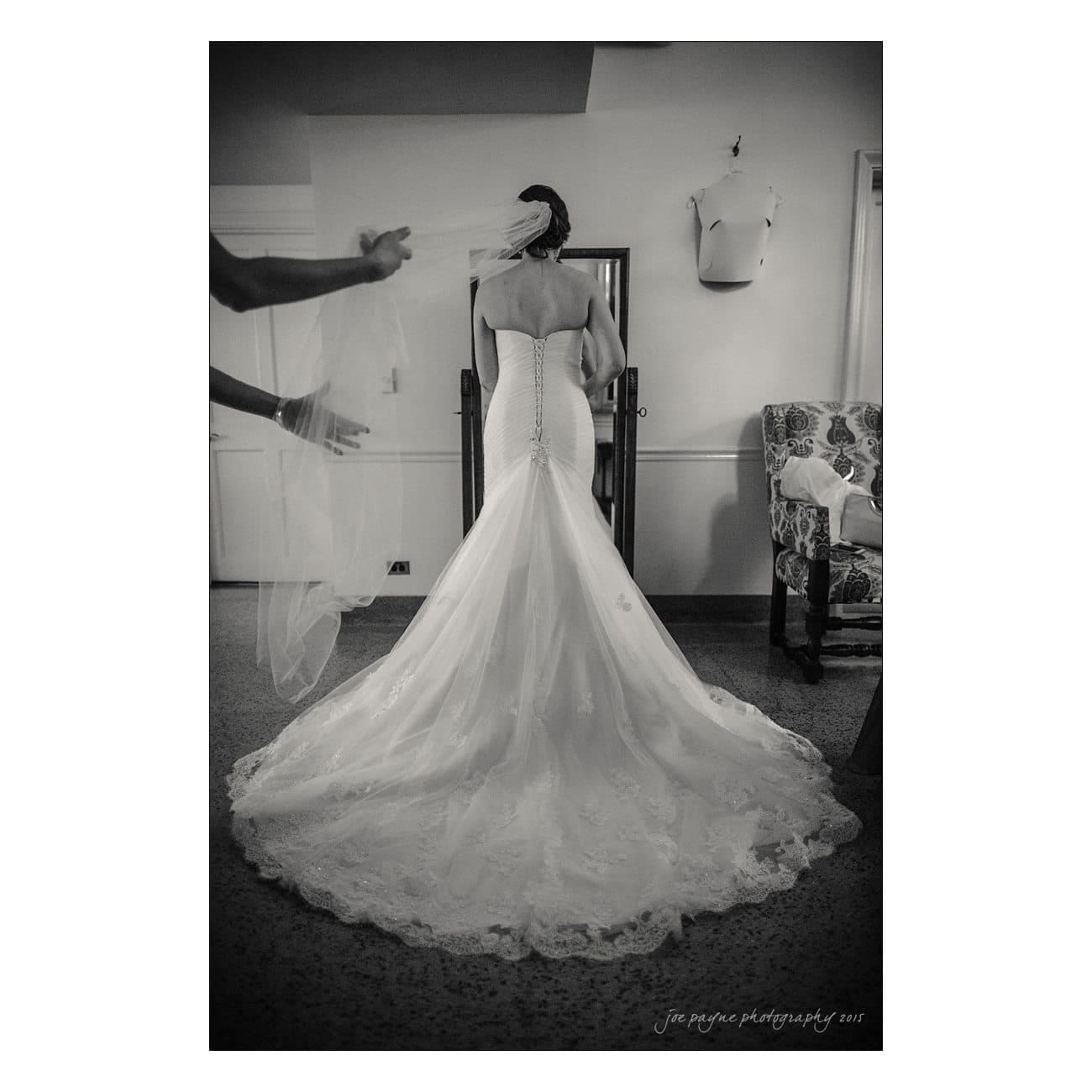 Bride in a long, elegant gown stands with her back to the camera. Her veil is being adjusted by another person. The dress features a long, flowing train. The setting is indoors with various furniture pieces around.