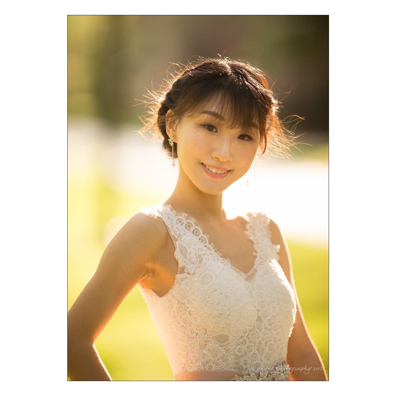 A young woman in a sleeveless lace dress stands outdoors, smiling softly. Her hair is styled up, and she is illuminated by warm, natural light, creating a serene and graceful ambiance.