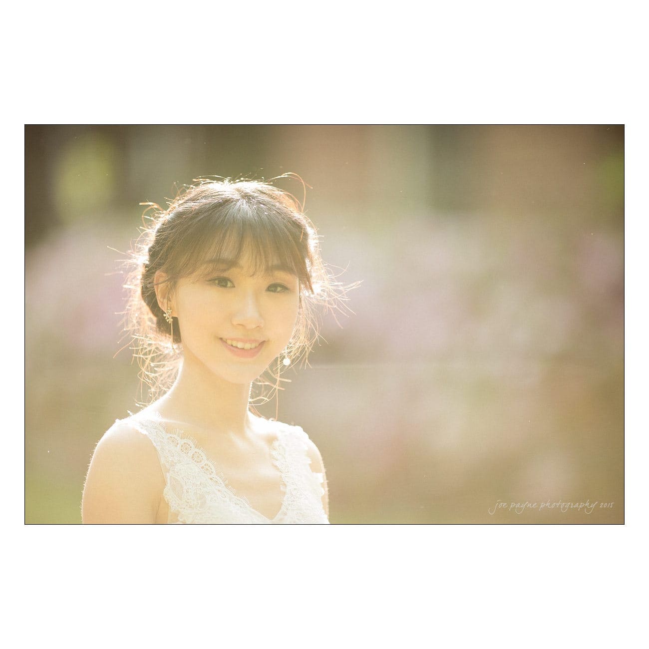 A woman in a lace dress smiles warmly while standing outdoors. Sunlight creates a soft, glowing effect around her. The background is softly blurred, enhancing the serene and dreamy atmosphere.