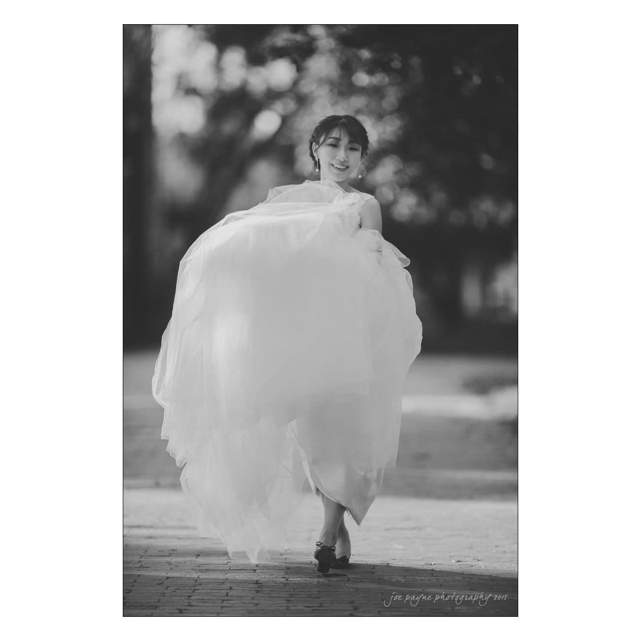 A black and white image of a person in a flowing wedding dress walking outdoors. The background features blurred trees and a pathway. The person appears joyful and is holding the dress slightly lifted.