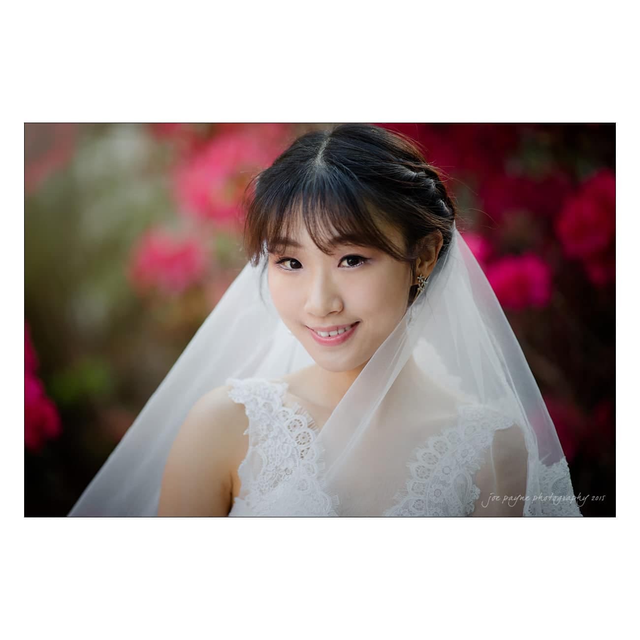 A bride smiles warmly, wearing a white lace dress and veil. Vibrant red flowers provide a colorful background.