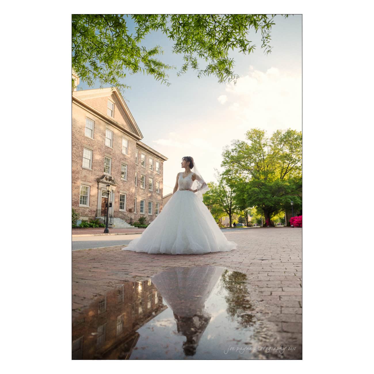 A bride in a white gown stands on a brick pathway beside a historic building. Trees surround the area, and her reflection is visible in a puddle. The sky is partly cloudy.